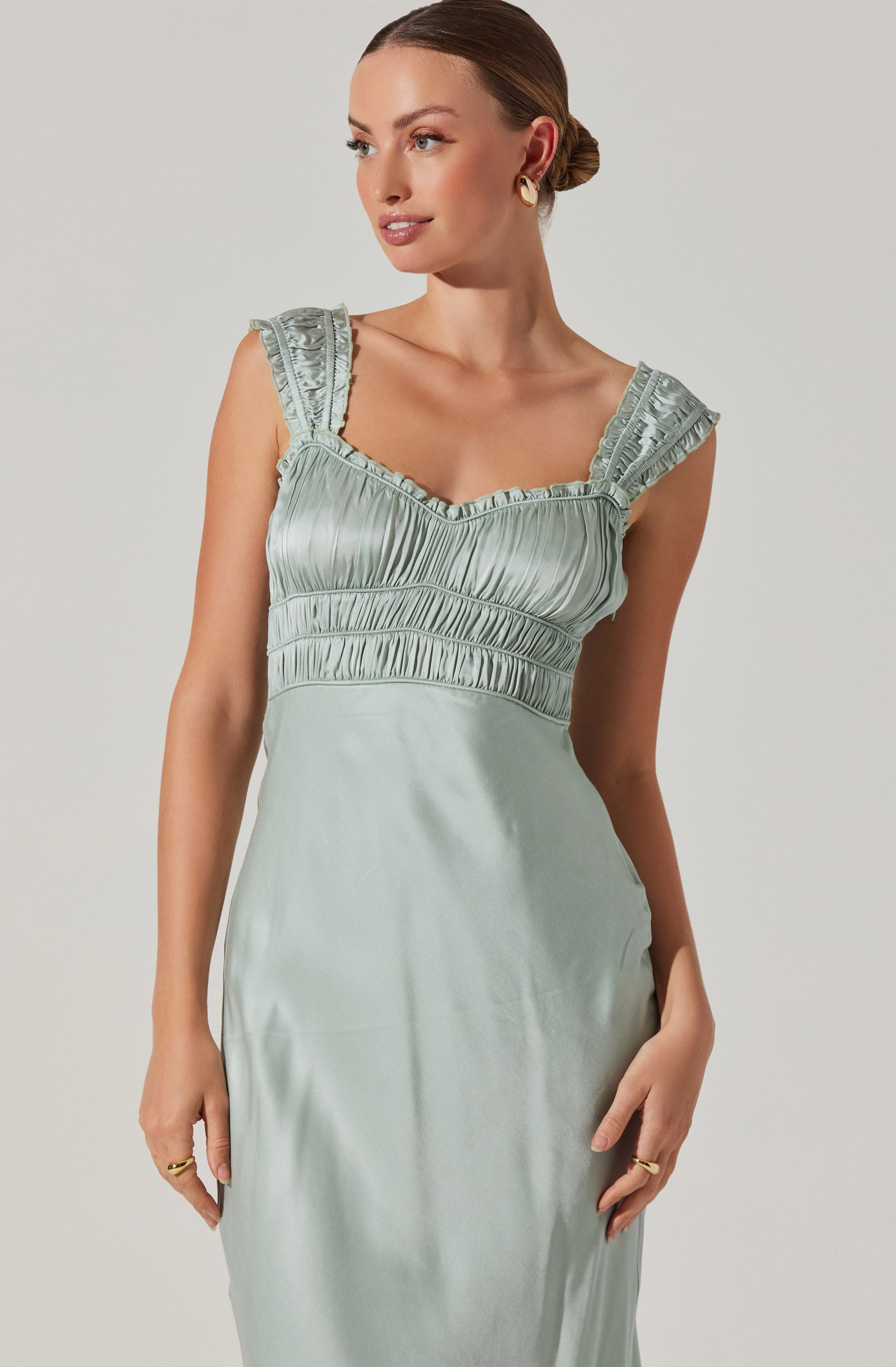 A woman is wearing a sleeveless, light-colored satin dress with ruched detailing on the bodice and ruffled shoulder straps.