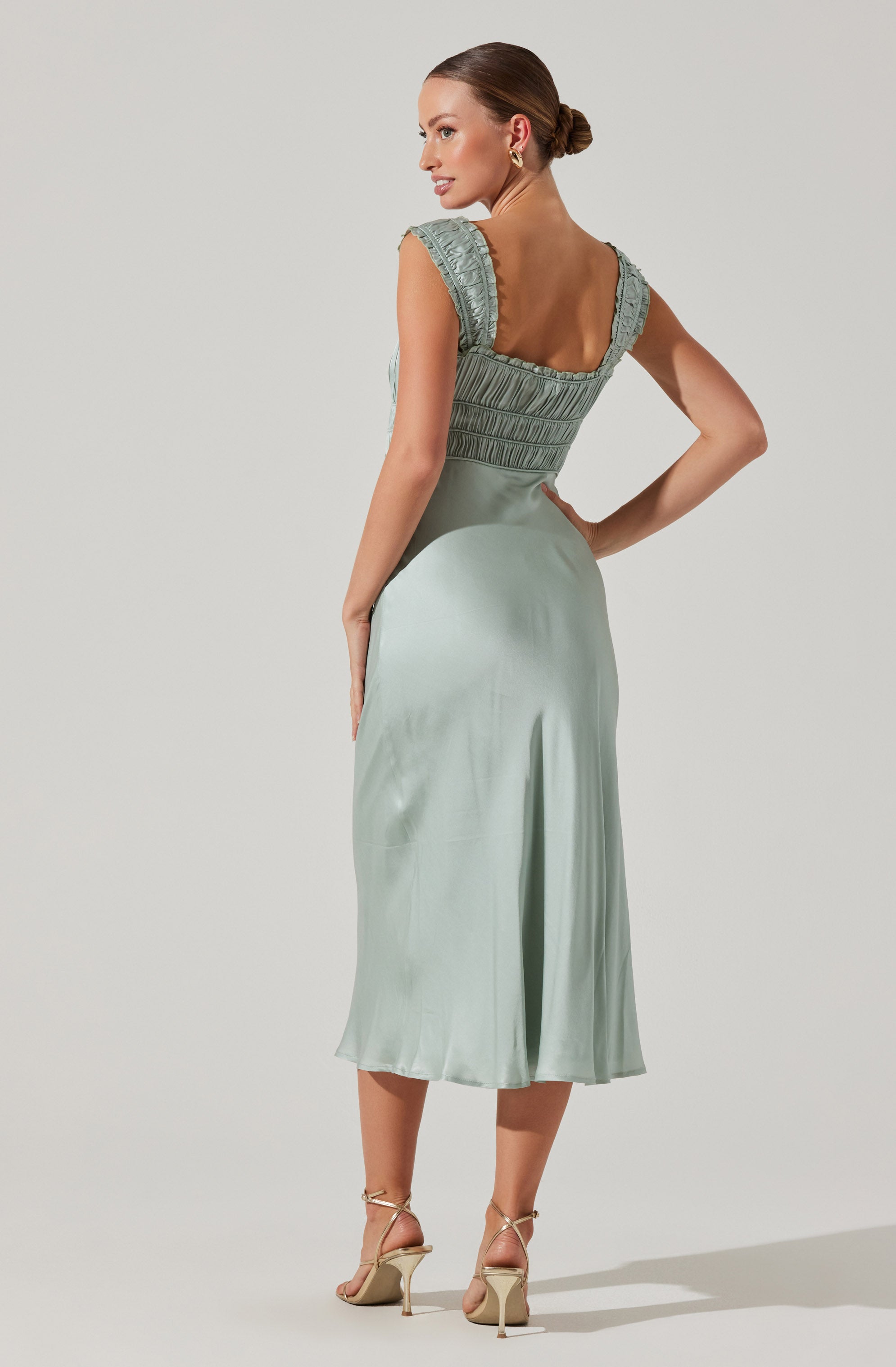 A woman is wearing a sleeveless, light green satin dress with a fitted bodice featuring intricate smocking and ruffle details on the shoulders. The dress has a square neckline at the back and a flowing midi length skirt. She is also wearing beige high-heeled sandals with ankle straps. Her hair is styled in a sleek bun, and she is wearing small gold hoop earrings.