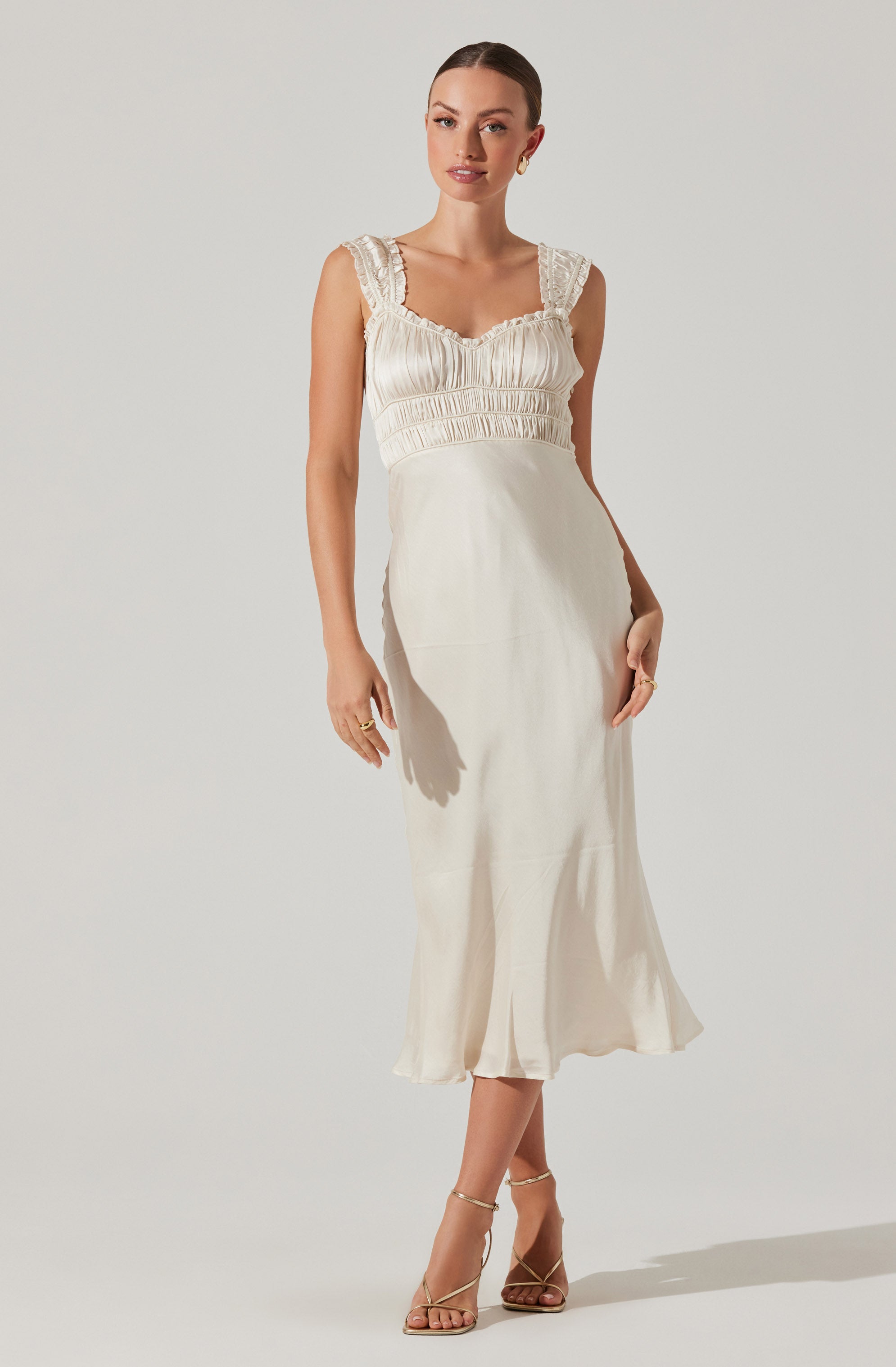 A woman is wearing a sleeveless, cream-colored satin dress with ruffled shoulder straps, a ruched bodice with a sweetheart neckline, and a fitted waist. The skirt is long and flowing, reaching mid-calf. She is wearing strappy high-heeled sandals and small hoop earrings.