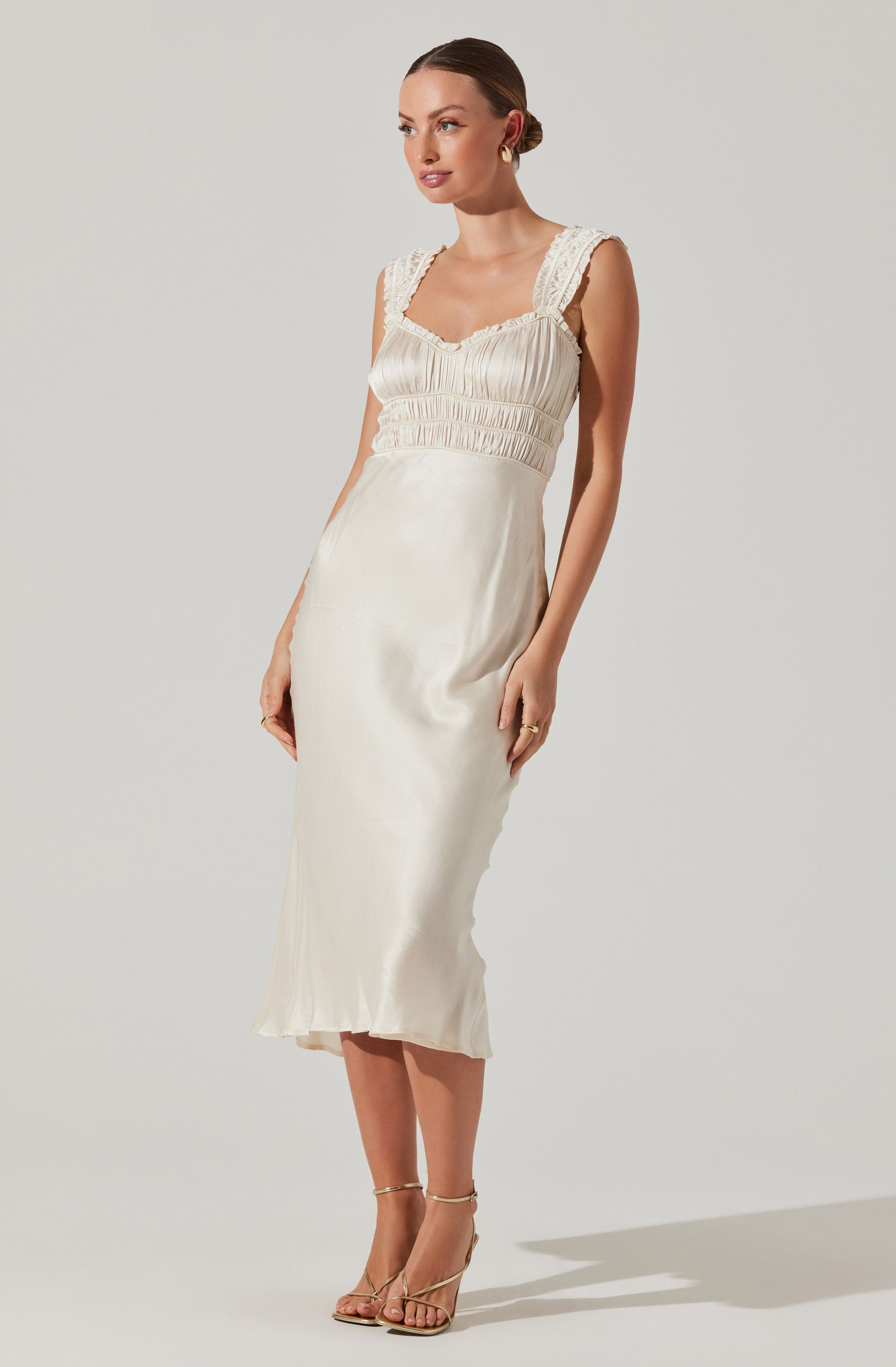 A woman is wearing a sleeveless, cream-colored satin dress with lace shoulder straps. The dress features a fitted bodice with ruched detail and an empire waist, flowing into a midi-length skirt. She is also wearing nude high-heeled sandals with ankle straps.