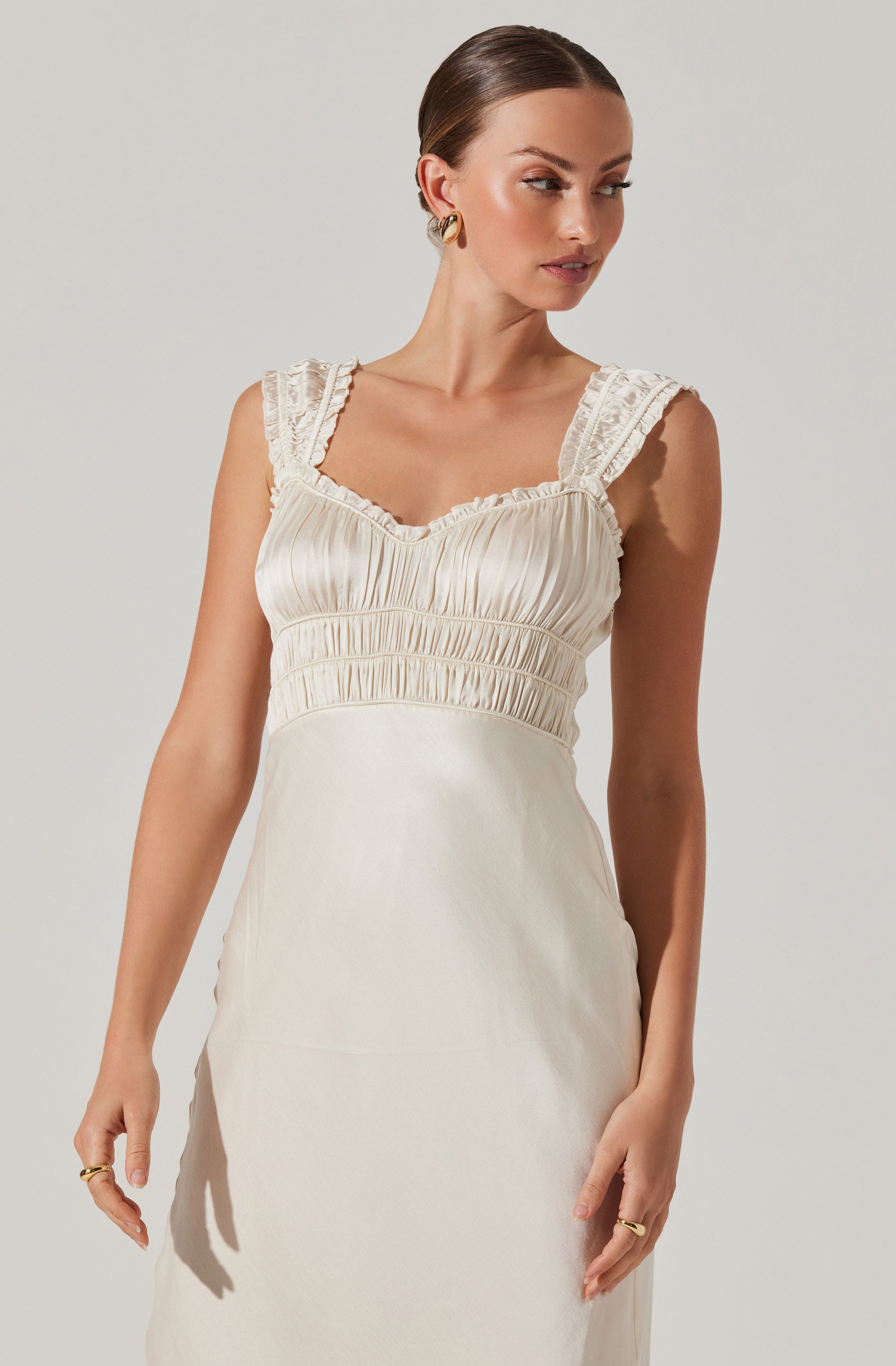 A woman is wearing a sleeveless, cream-colored dress with ruched detailing on the bodice, ruffled shoulder straps, and a smooth, flowing skirt.