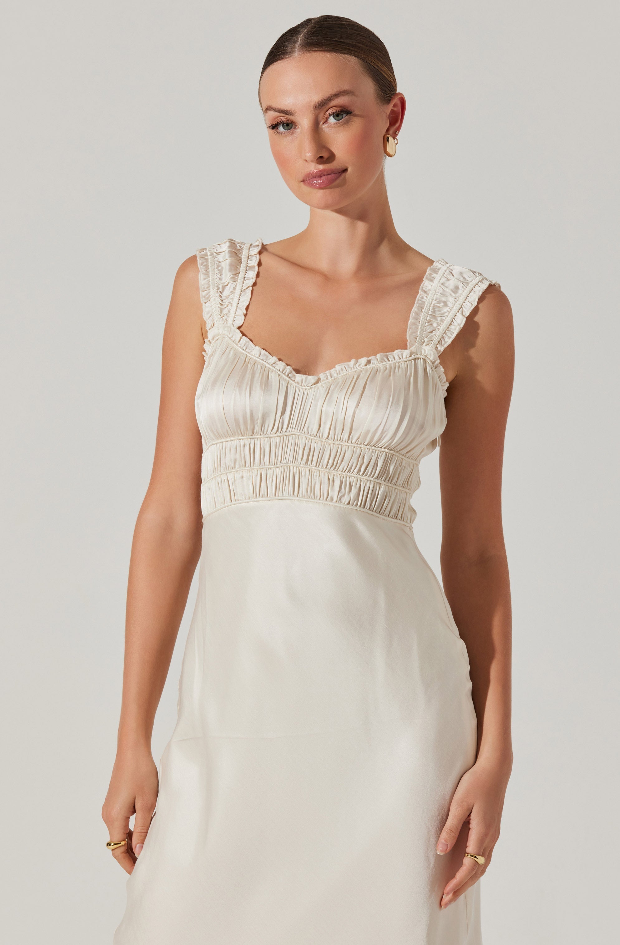 A woman is wearing a sleeveless, cream-colored dress with ruffled lace shoulder straps and a fitted bodice featuring horizontal ruching. The dress appears to be made of a silky material with a subtle sheen.
