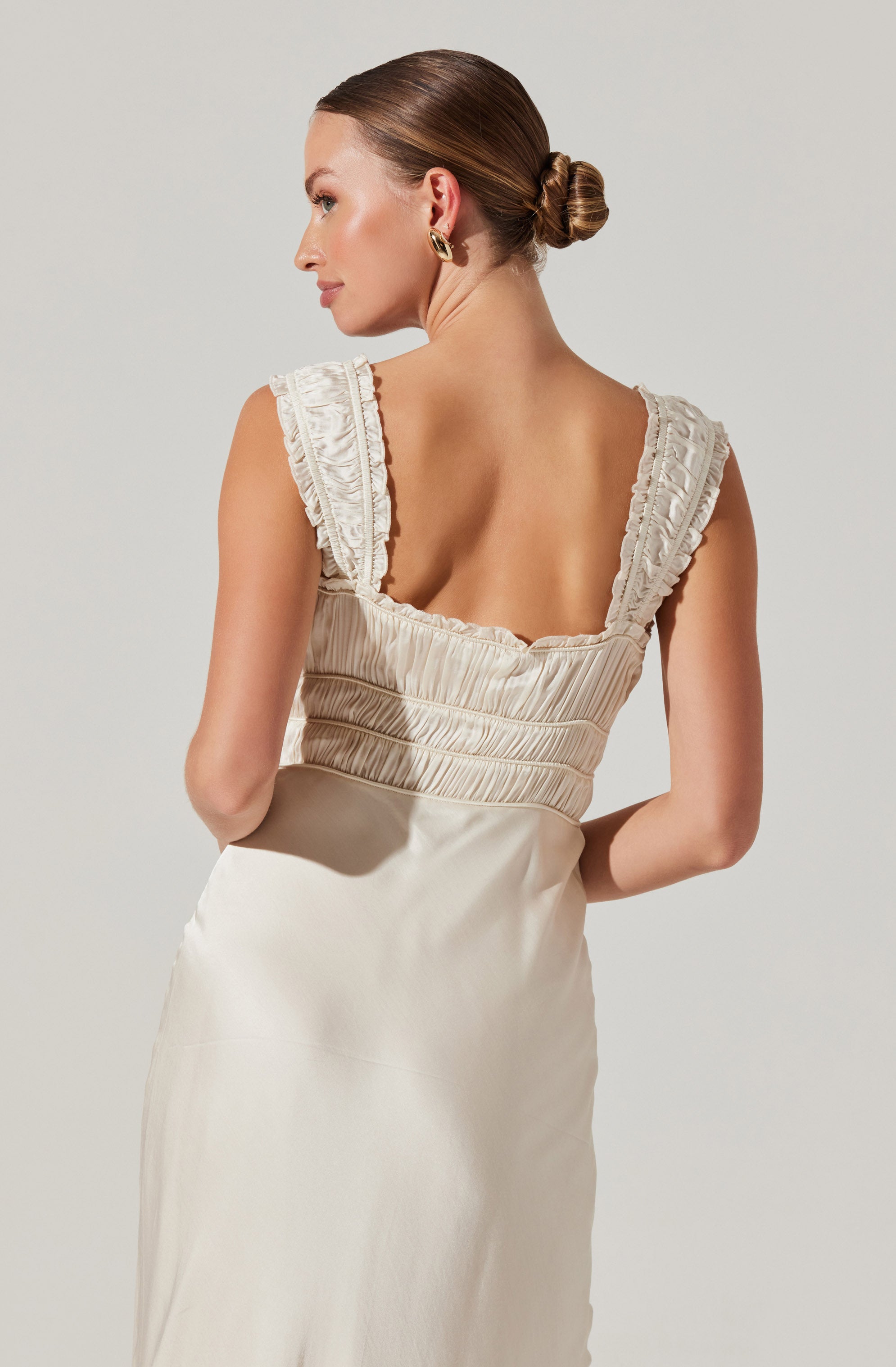 A woman wearing a sleeveless, cream-colored dress with ruffled straps and ruched detailing on the bodice. The dress has a fitted waist and flows down in a smooth fabric.