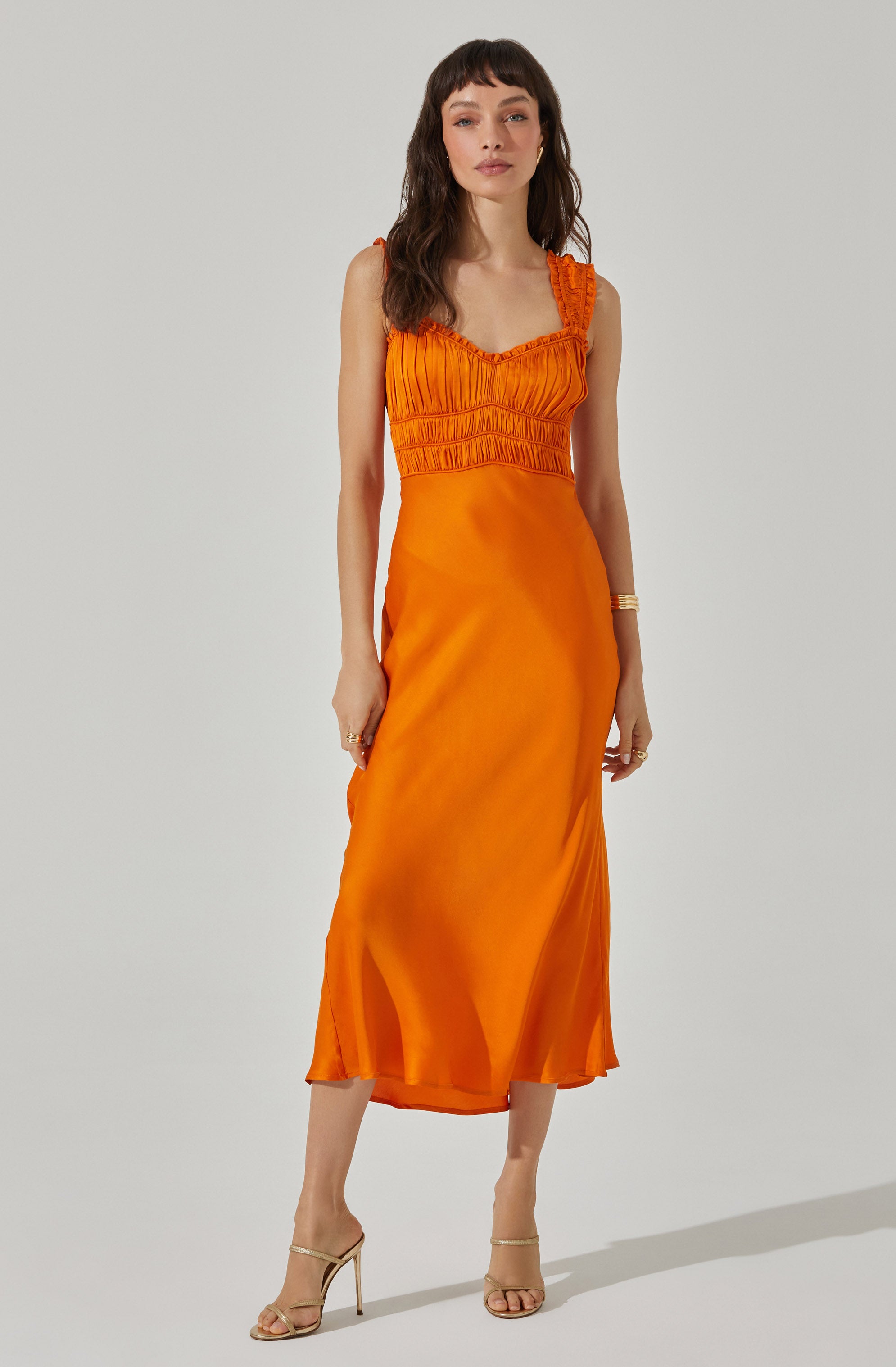 A woman is wearing a sleeveless, vibrant orange dress with a fitted bodice featuring gathered and ruched fabric details along the top and shoulder straps. The dress has a smooth, flowing skirt that reaches mid-calf length, made of a shiny satin-like material. She is accessorized with gold jewelry, including earrings, rings, and bangles, and is wearing strappy high-heeled sandals.