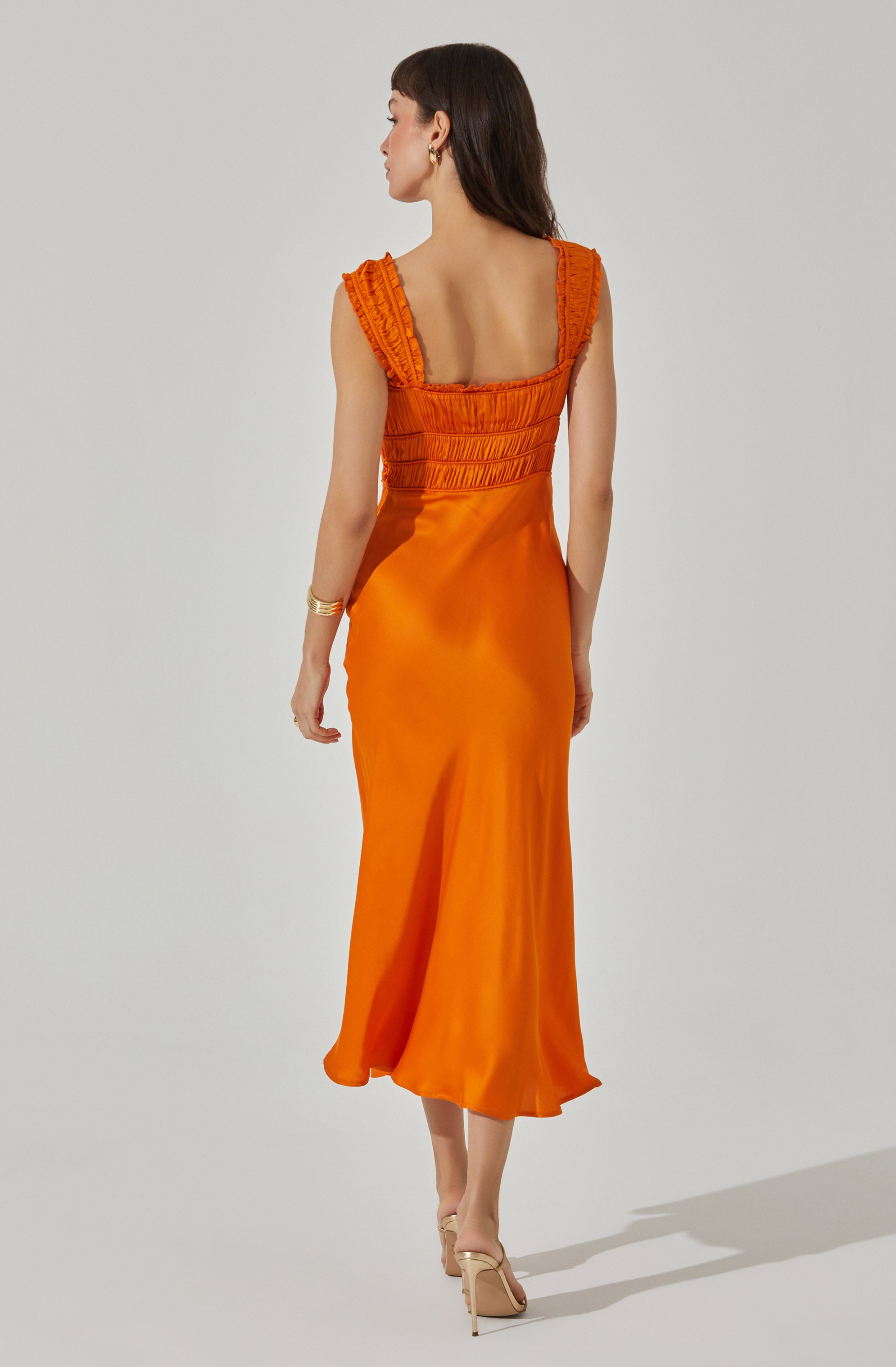 A woman is wearing a sleeveless, fitted, bright orange satin dress with gathered detailing on the bodice and ruffled straps. The dress has a smooth, shiny texture and flows down to an asymmetrical hemline that falls below the knees. She is also wearing nude high-heeled sandals.