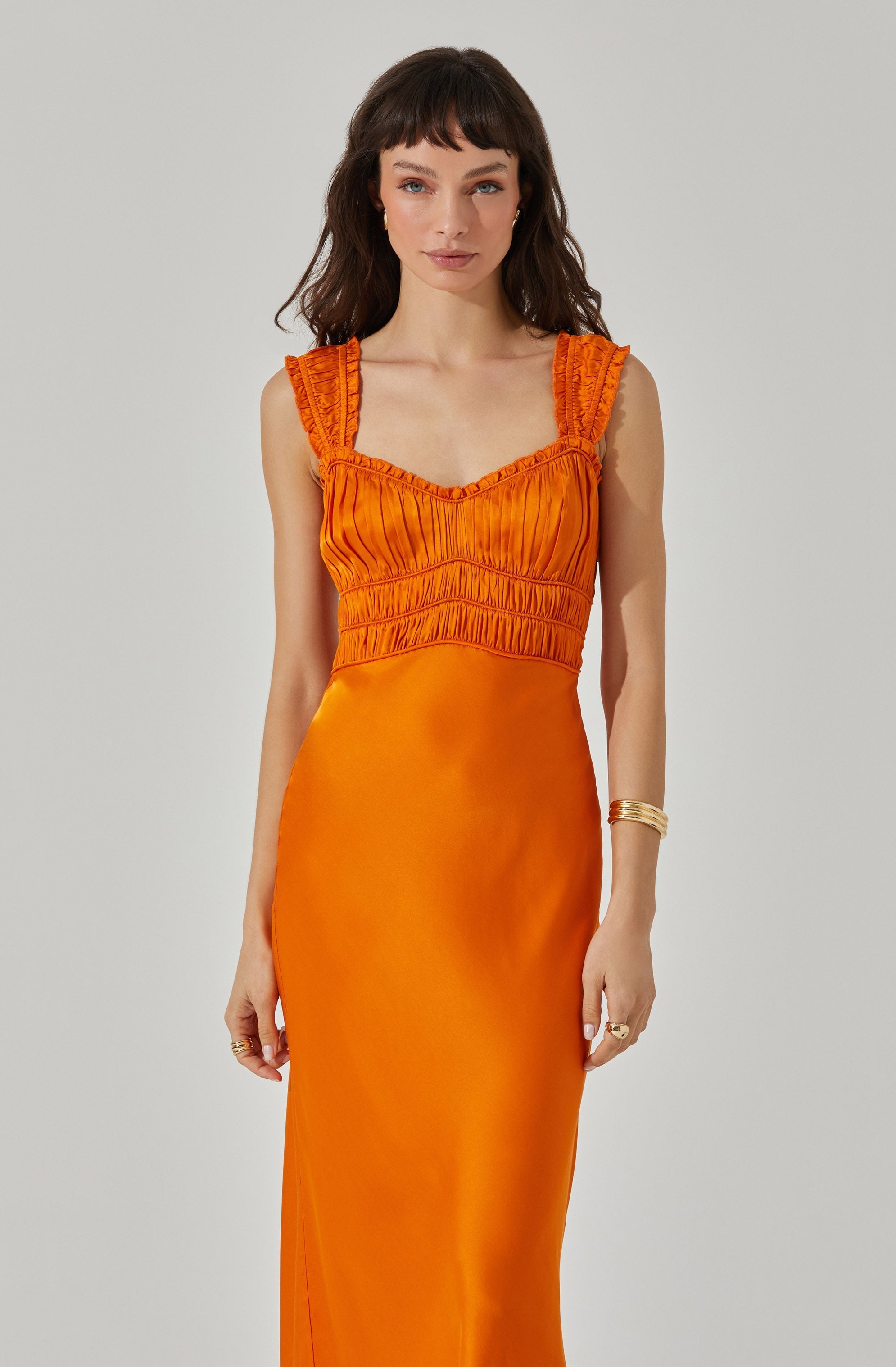 A woman is wearing a sleeveless, bright orange dress with ruched details on the bodice and ruffled shoulder straps. The dress has a fitted waist and a long, smooth skirt that extends to the floor. She has accessorized with gold bangles on her right wrist and rings on her left hand.