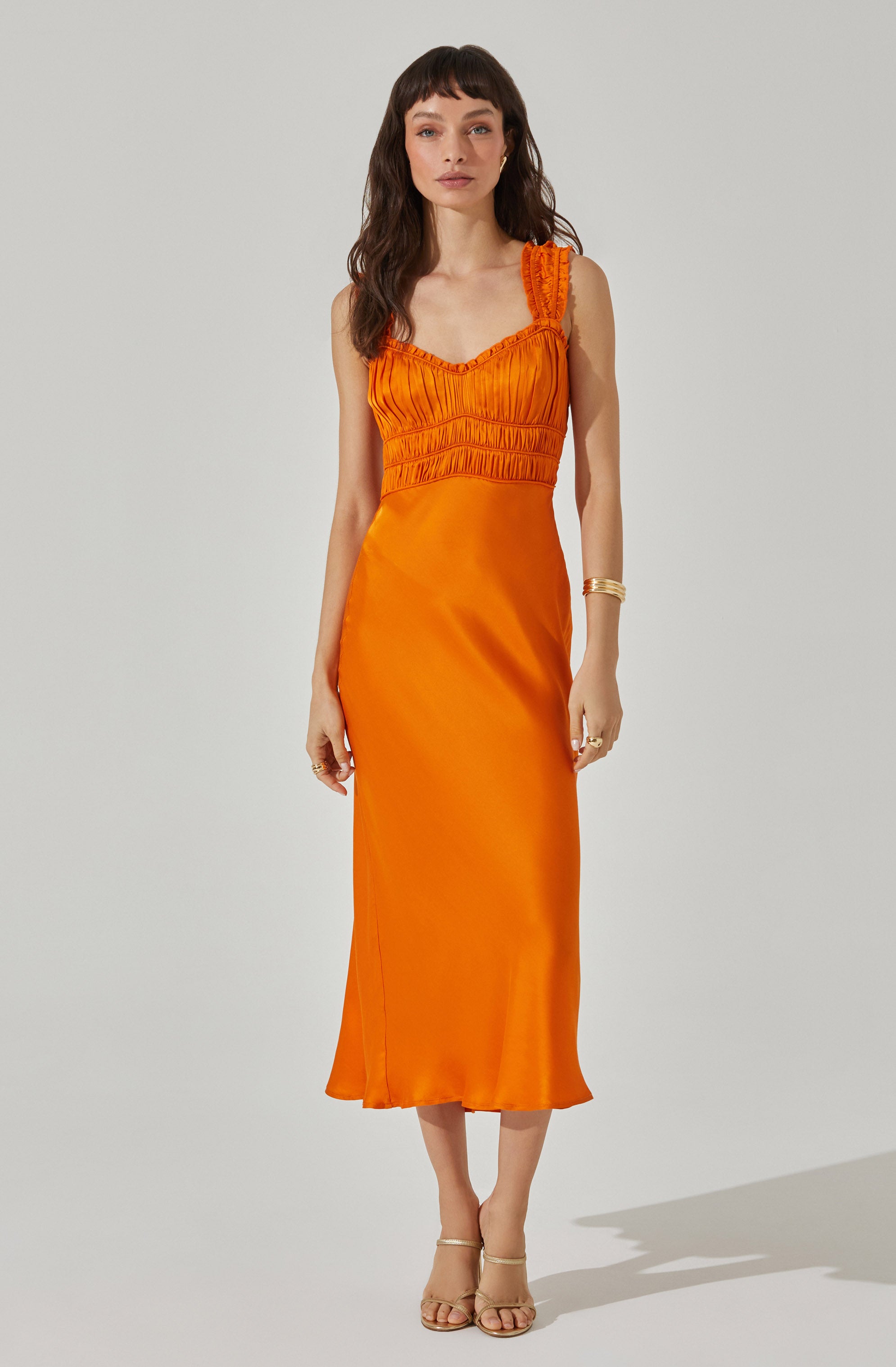 A woman is wearing an orange sleeveless dress with a fitted bodice featuring vertical pleats and shirred details, and a flowy, ankle-length skirt made of shiny satin-like fabric. She has paired the dress with strappy nude heels and some gold jewelry on her arm and fingers.