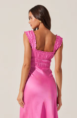 A woman is wearing a sleeveless, bright pink satin dress with ruffled shoulder straps. The top of the dress features gathered fabric with a ruched appearance, and the fabric extends smoothly down to the skirt. She has long, dark brown hair and is wearing gold earrings.