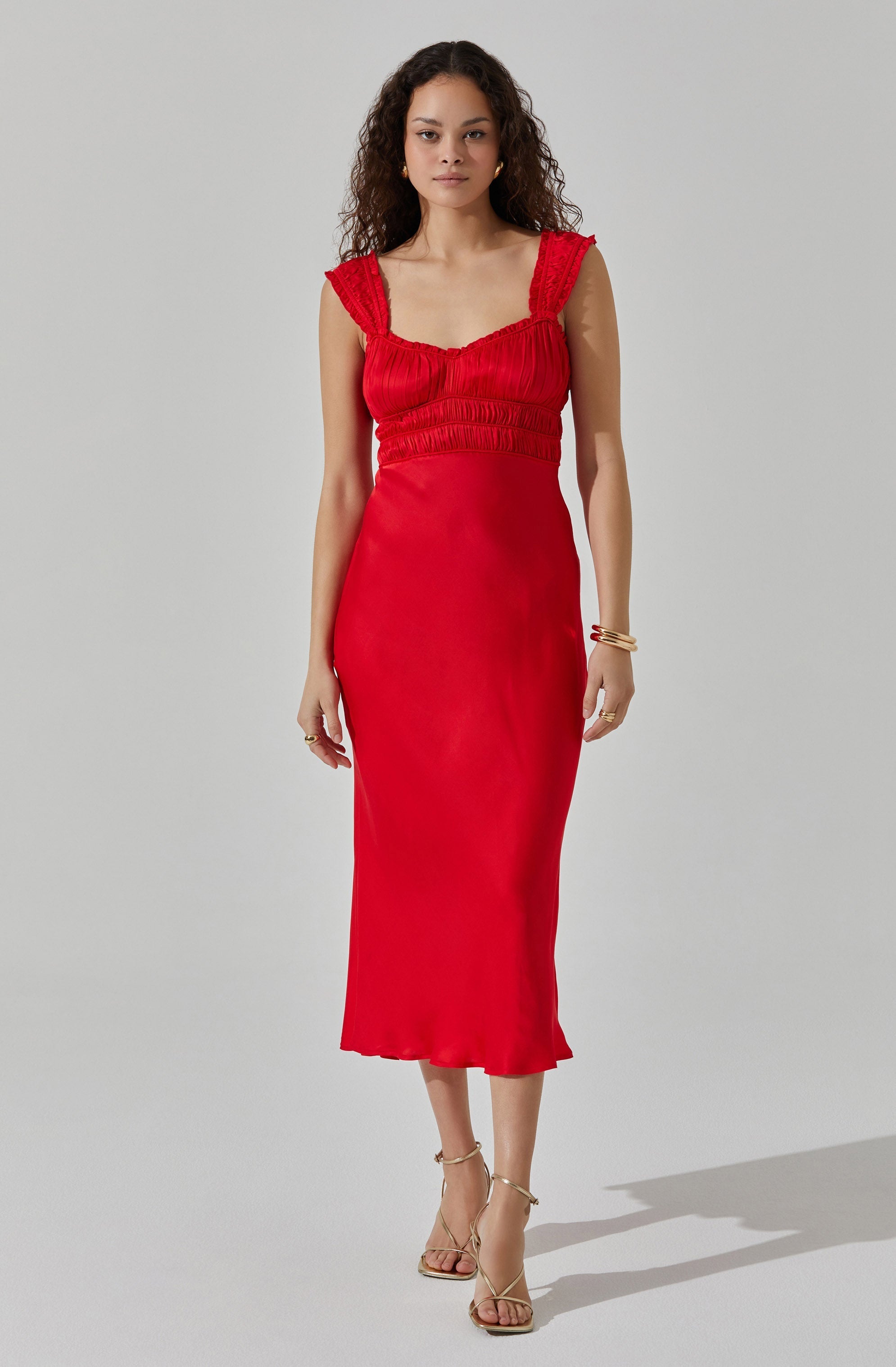 A woman is wearing a sleeveless, fitted, red dress with ruffle details on the shoulder straps, a ruched bodice, and a smooth, flowing skirt that reaches mid-calf. She is accessorized with gold jewelry and strappy gold high-heeled sandals.