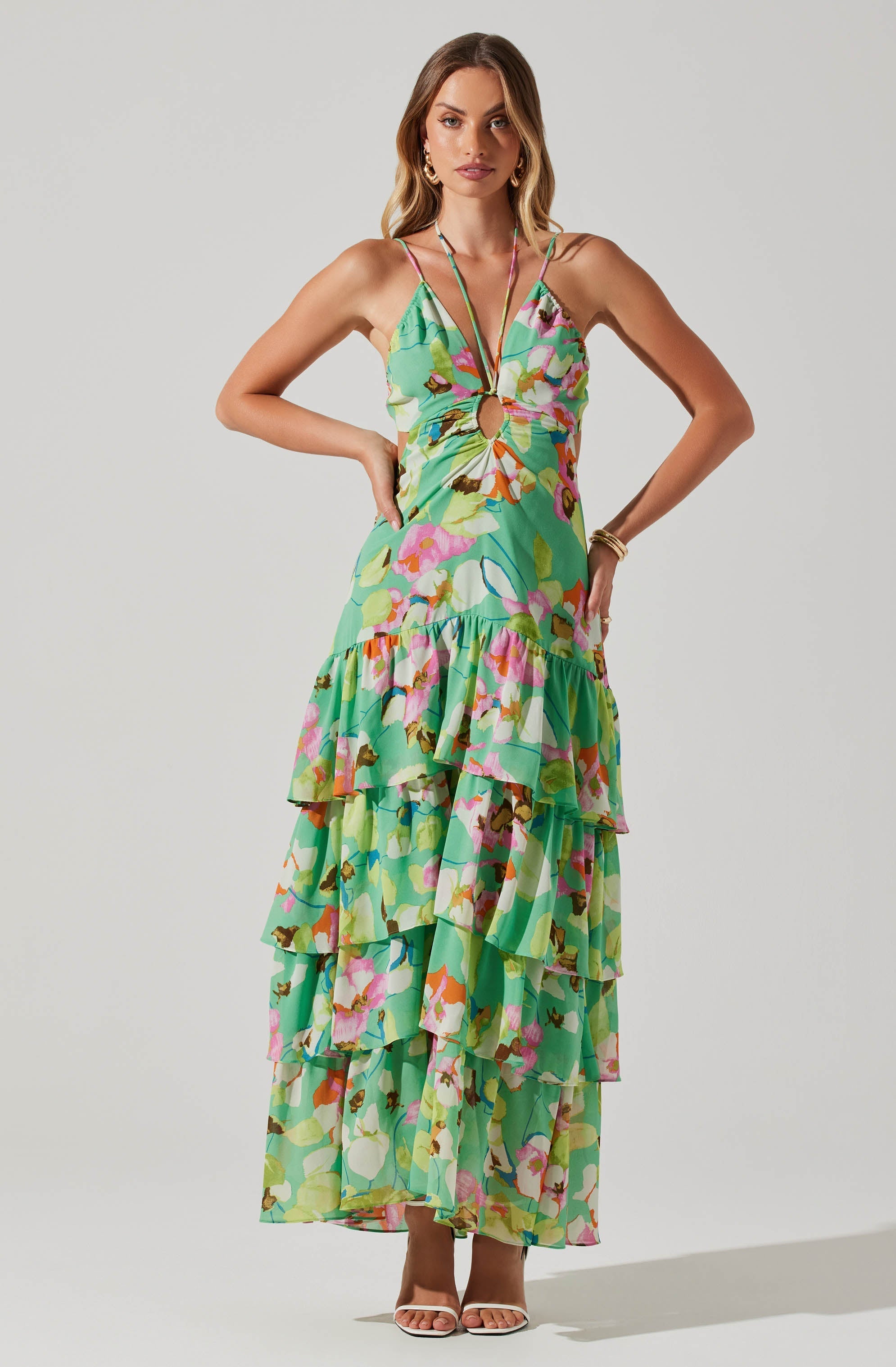 A woman is wearing a long, colorful, floral-print dress with a tiered, ruffled skirt. The dress features thin shoulder straps and a large circular cutout at the front, with the fabric gathered around the cutout. The dress has a bright pattern with shades of green, pink, orange, and white. She is accessorized with gold hoop earrings, a gold bracelet, and a necklace with multiple strands. She is wearing open-toed high-heeled sandals.