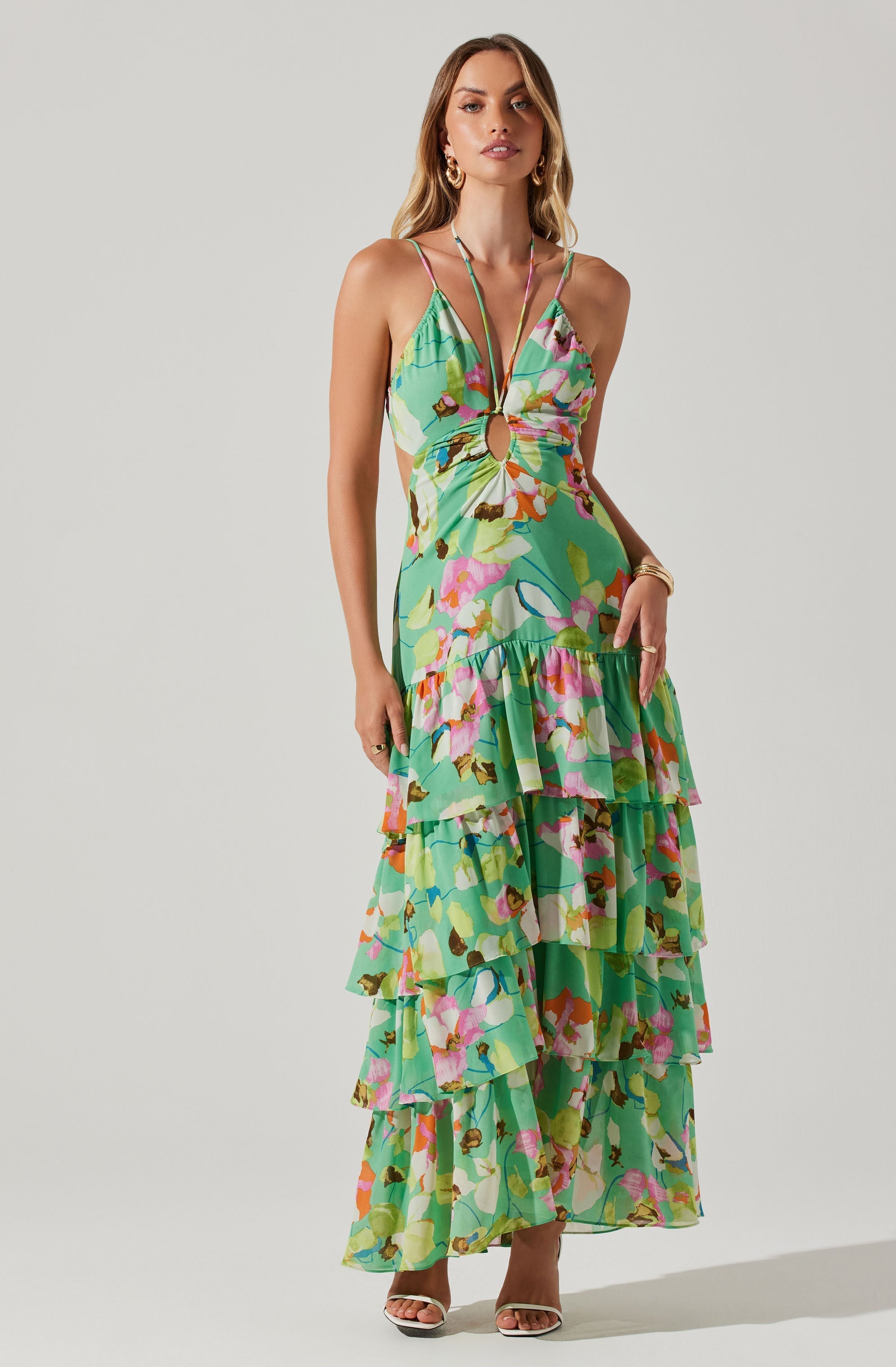 A woman is wearing a colorful, sleeveless maxi dress with thin spaghetti straps. The dress features a vibrant, multicolored floral and abstract pattern with shades of green, pink, purple, orange, and brown. It has a gathered, ruffled tiered skirt that extends to her ankles and a circular cut-out detail at the front connected by a small ring.