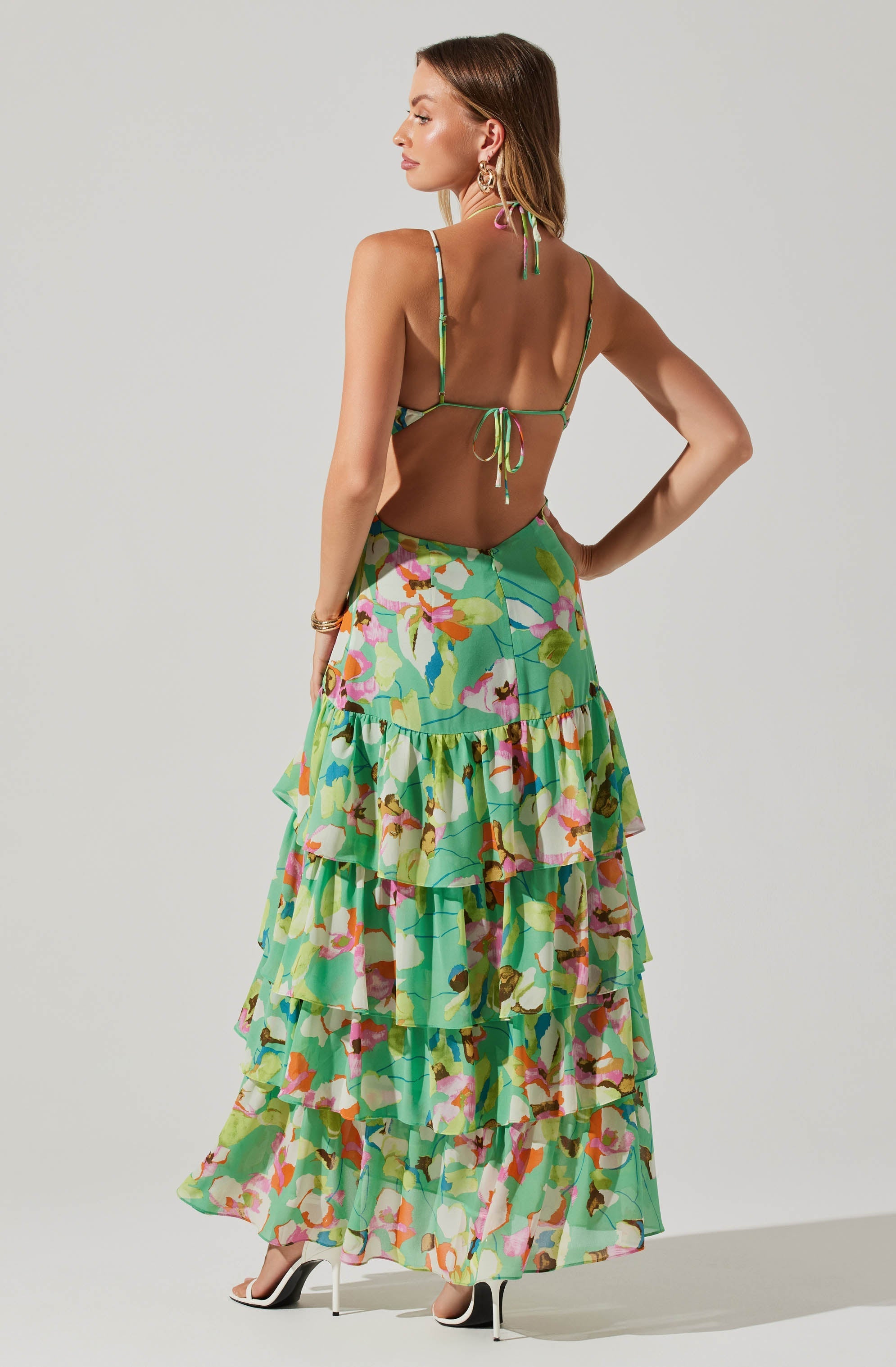 A woman is wearing a colorful, floral-printed outfit consisting of a thin-strap, backless crop top with ties at the neck and back, paired with a long, tiered skirt featuring a vibrant, multicolored floral pattern.