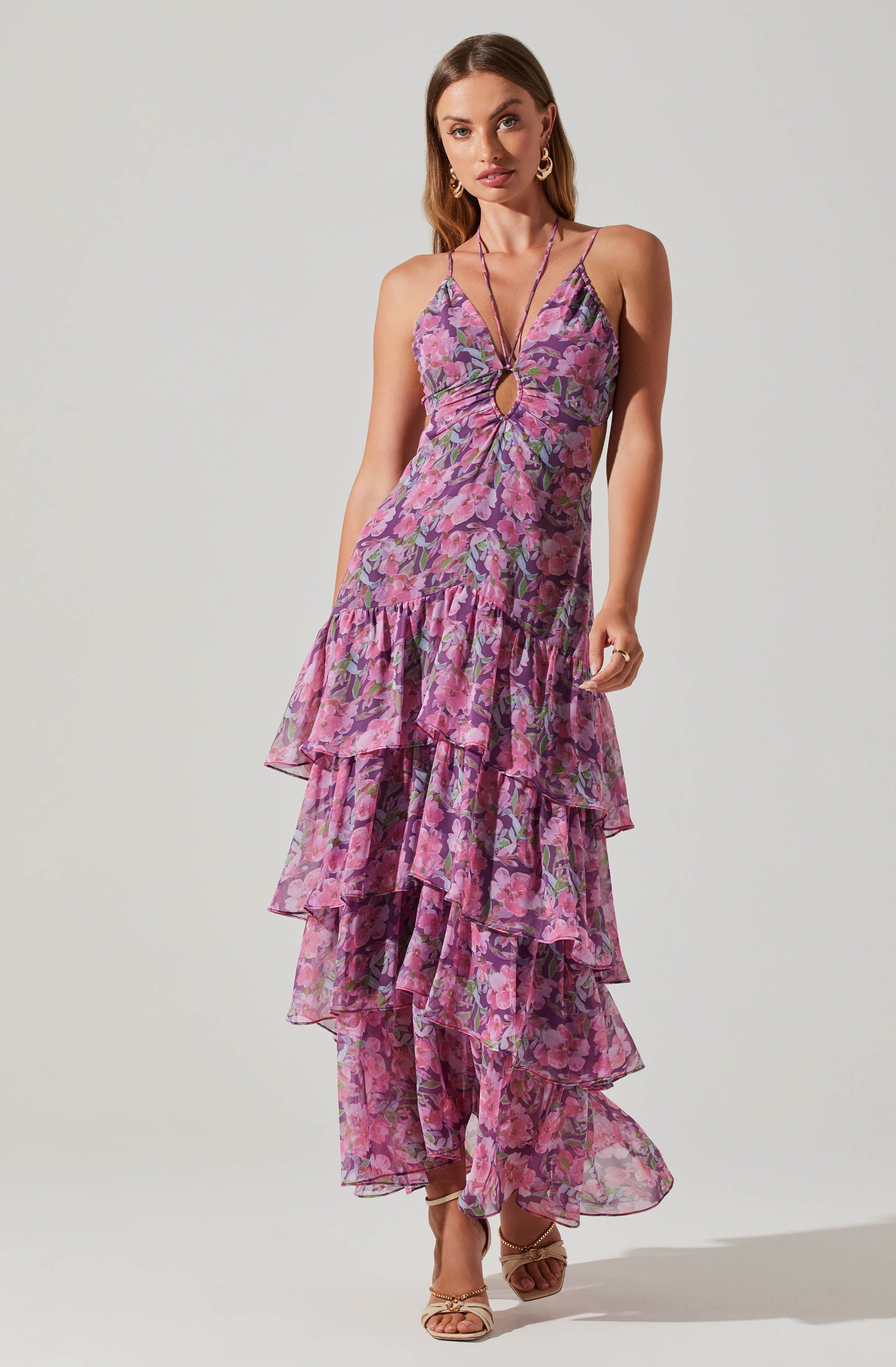 A woman is wearing a sleeveless, v-neck maxi dress with thin spaghetti straps. The dress features a floral pattern with shades of pink, purple, and green. It has a keyhole cutout at the front just below the neckline and a ruffled, tiered design that extends from the waist to the hem. She accessorizes with gold hoop earrings and a delicate necklace, and is wearing open-toe heels.