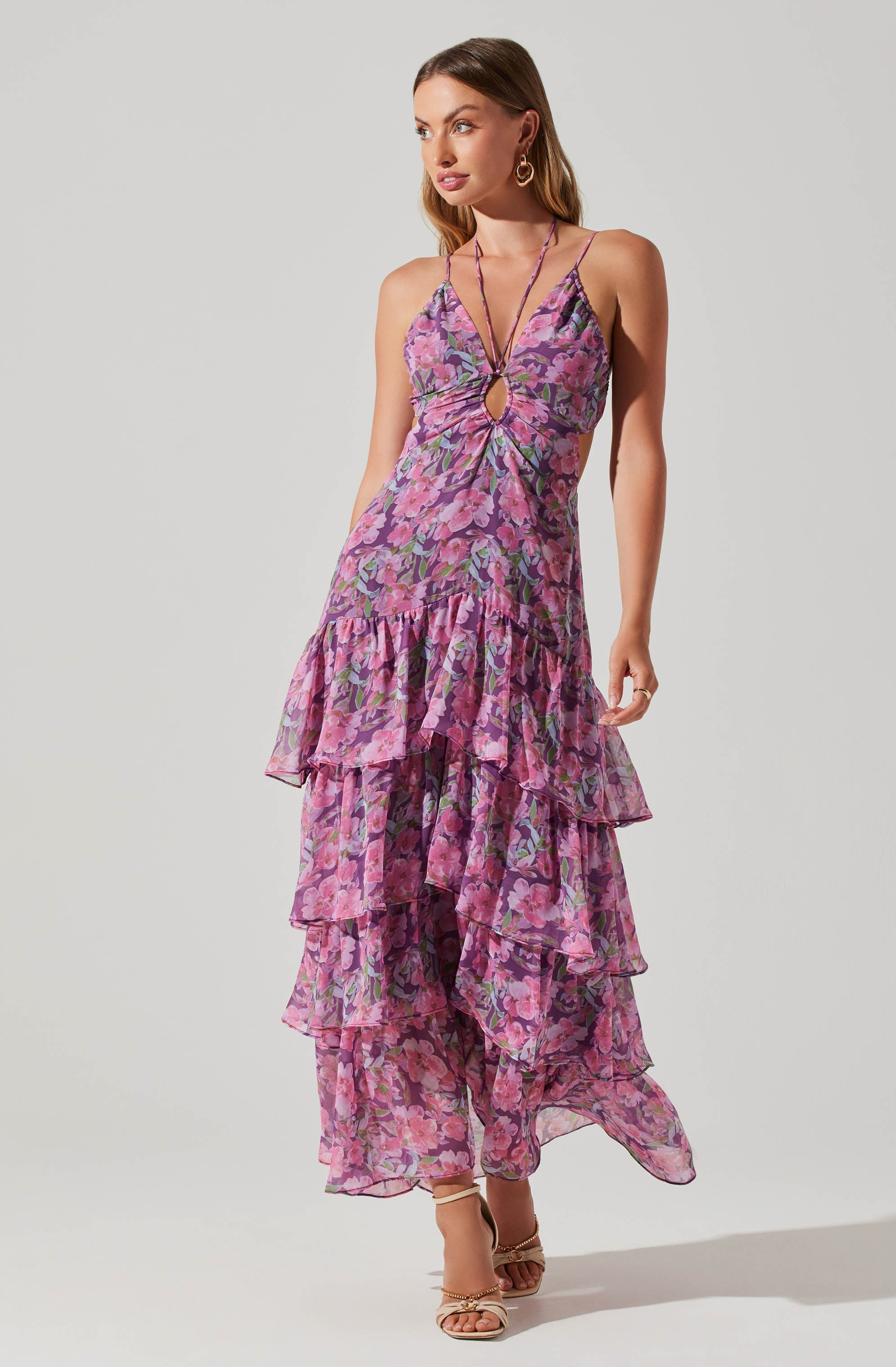 A woman is wearing a sleeveless, floral maxi dress with thin spaghetti straps and a keyhole cutout at the front. The dress features a tiered, ruffled skirt in shades of pink, purple, and green. She has on gold hoop earrings and nude high-heeled sandals.
