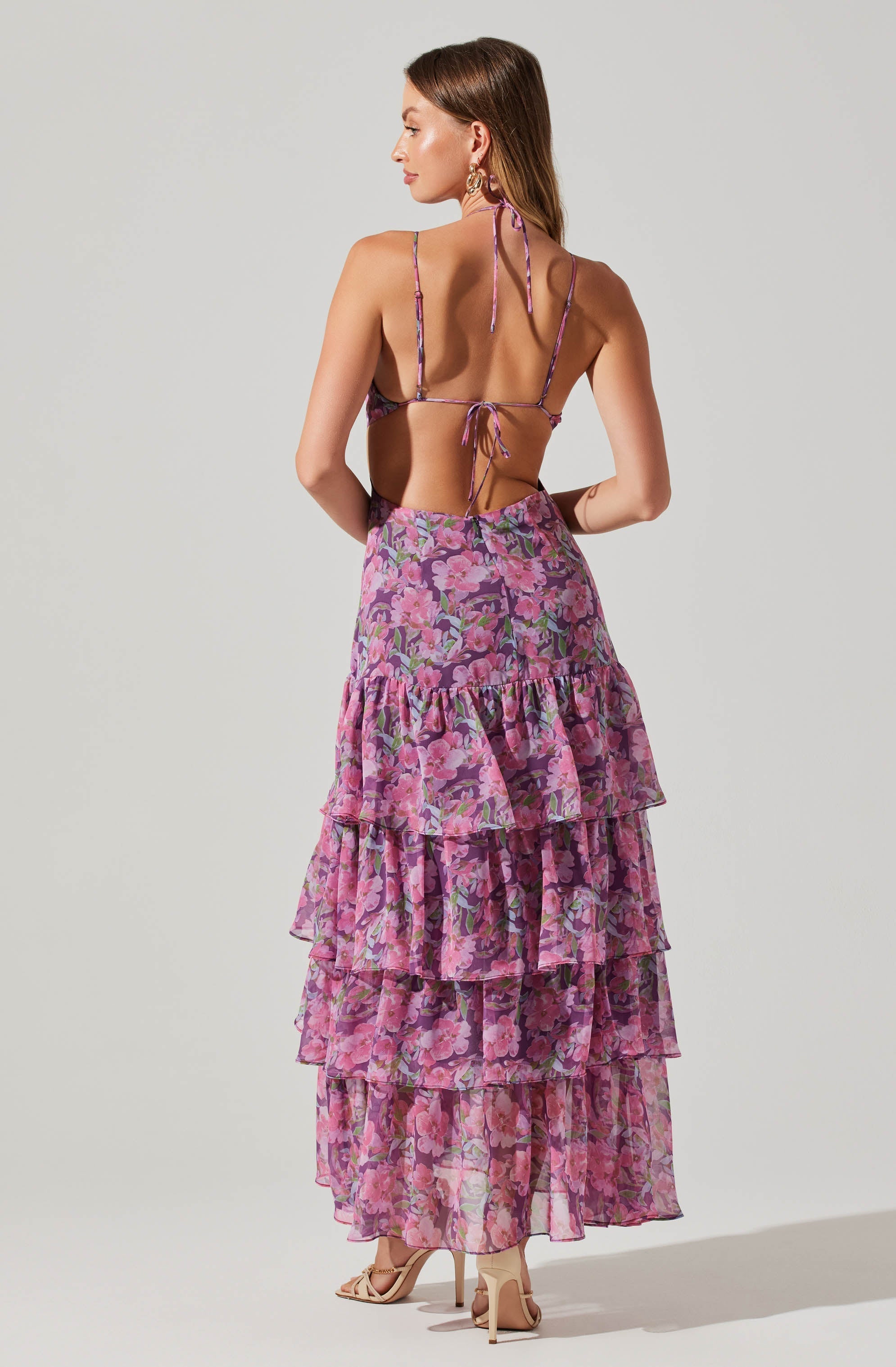 A woman is wearing a backless, purple and pink floral spaghetti strap top with tie closures at the neck and back. She is also wearing a long, flowing maxi skirt with multiple ruffled tiers in a matching pink and purple floral pattern. She has on high-heeled sandals and statement earrings.