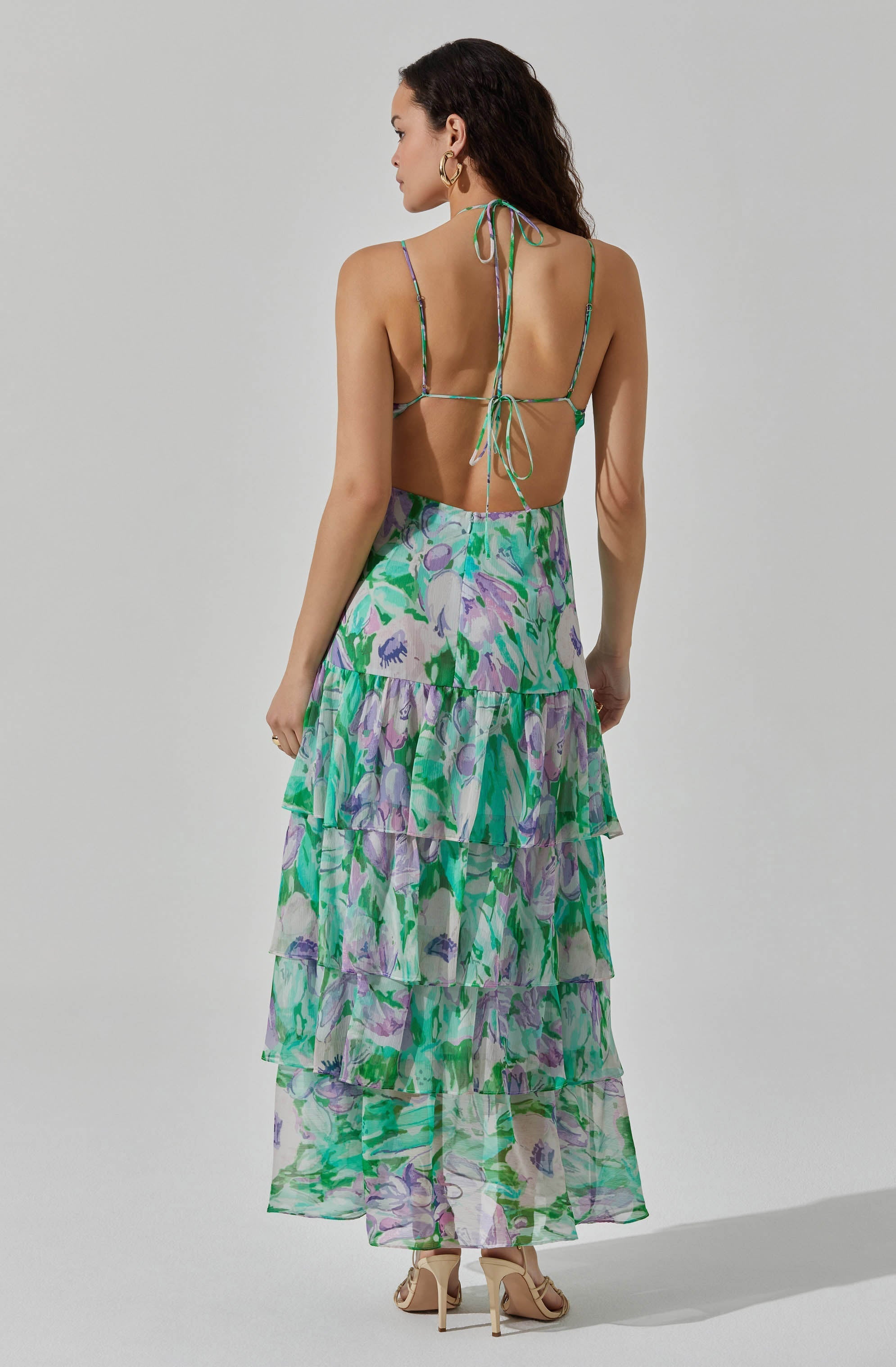 A woman is wearing a backless, strappy top with thin, multicolored straps tied into bows at the neck and back. She is also wearing a long, flowy skirt with a tiered design, featuring a vibrant floral print in shades of green, purple, and white.