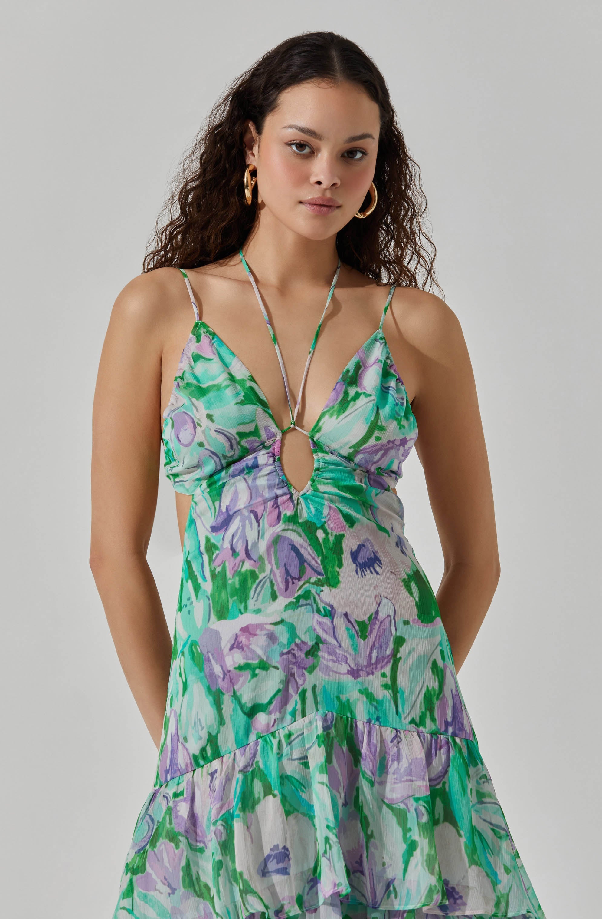 A woman is wearing a sleeveless, flowy dress with spaghetti straps that tie at the shoulders. The dress features a vivid, abstract floral pattern in shades of green, purple, blue, and white. The fabric appears lightweight and airy, suitable for warm weather. The dress has a keyhole cutout at the front, accented with a small metal ring connecting the straps. The skirt of the dress is tiered with ruffled layers, adding a playful, feminine touch.