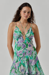 A woman is wearing a sleeveless, flowy dress with spaghetti straps that tie at the shoulders. The dress features a vivid, abstract floral pattern in shades of green, purple, blue, and white. The fabric appears lightweight and airy, suitable for warm weather. The dress has a keyhole cutout at the front, accented with a small metal ring connecting the straps. The skirt of the dress is tiered with ruffled layers, adding a playful, feminine touch.