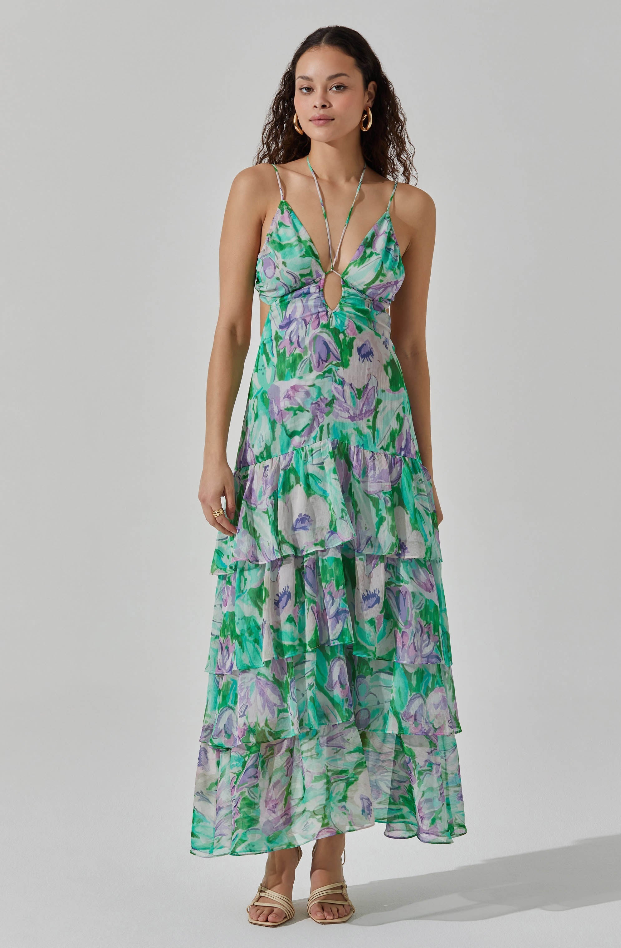 A woman is wearing a long, flowy dress with multiple tiers featuring a vibrant, abstract floral pattern in shades of green, purple, and white. The dress has thin spaghetti straps and a keyhole cutout at the front. She accessorizes with gold hoop earrings and a delicate necklace, and she is wearing open-toed, beige high-heeled sandals.