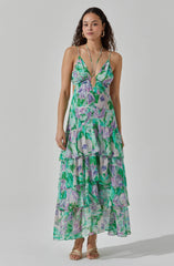 A woman is wearing a long, flowy dress with multiple tiers featuring a vibrant, abstract floral pattern in shades of green, purple, and white. The dress has thin spaghetti straps and a keyhole cutout at the front. She accessorizes with gold hoop earrings and a delicate necklace, and she is wearing open-toed, beige high-heeled sandals.