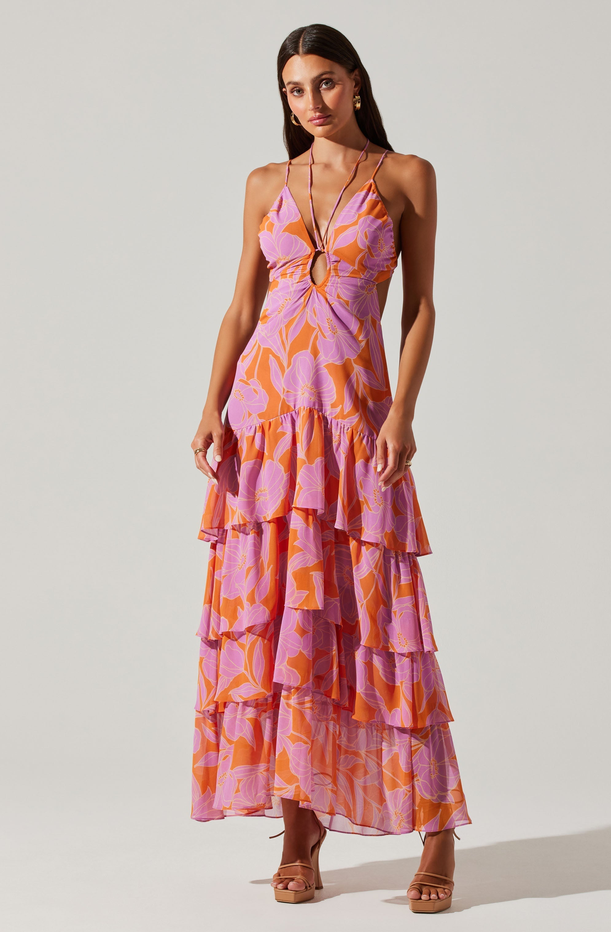 A woman is wearing a sleeveless, maxi dress with thin spaghetti straps. The dress features a vibrant floral pattern in shades of pink, purple, and orange. It has a deep V-neckline with a small circular cut-out detail at the center of the bust. The skirt has multiple layered ruffles that create a tiered effect, extending to the floor. She is also wearing nude-colored high-heeled sandals and has long, dark hair styled straight.