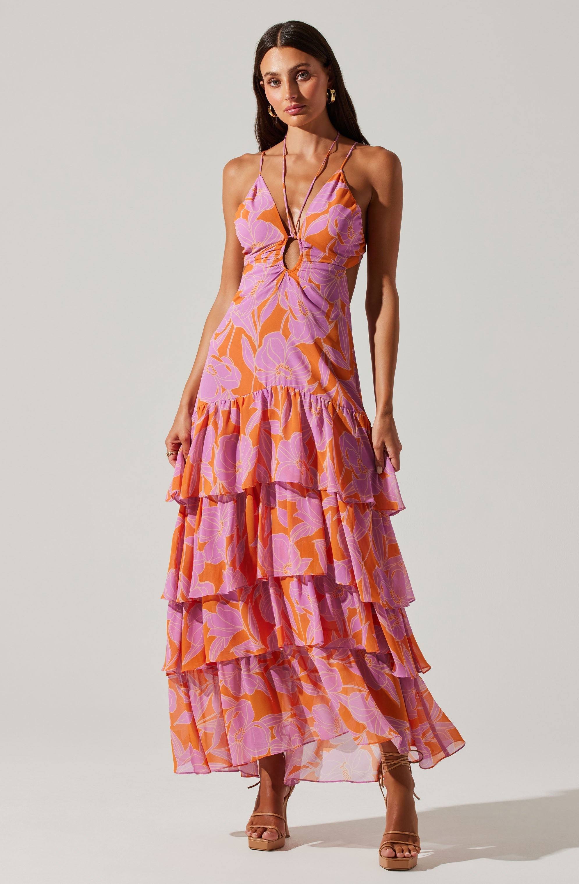 A woman is wearing a sleeveless, maxi dress with thin spaghetti straps. The dress features a vibrant floral pattern in shades of pink, purple, and orange. It has a fitted bodice with a keyhole cutout detail at the front, and a tiered, ruffled skirt that extends to the floor. The dress has a flowing, lightweight fabric, creating a summery and elegant look.