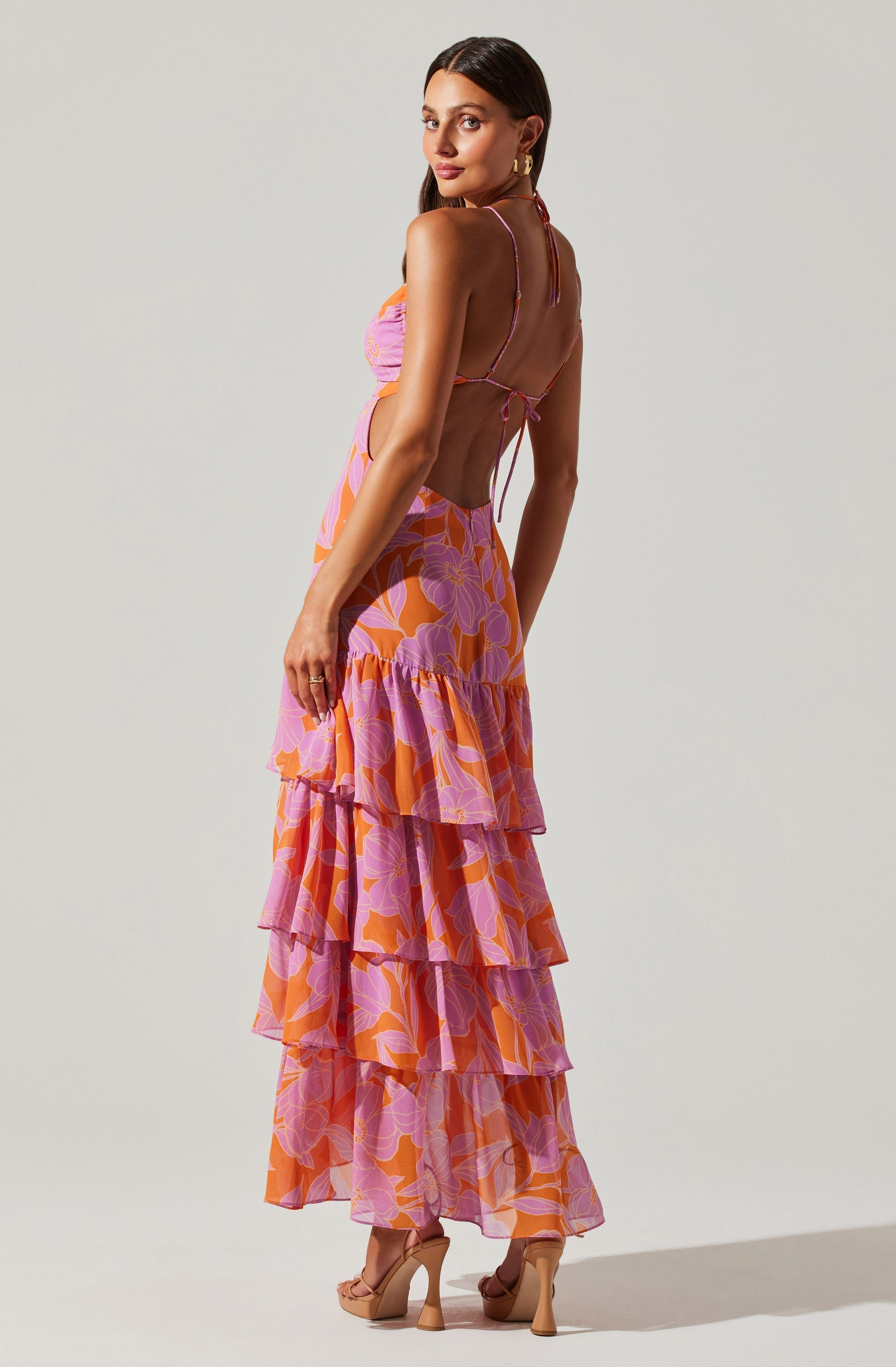 A woman is wearing a colorful two-piece outfit featuring a lavender and pink floral crop top with thin spaghetti straps and a matching long, tiered skirt with ruffled layers in pink, orange, and lavender floral patterns. She is also wearing tan high-heeled sandals and gold hoop earrings.