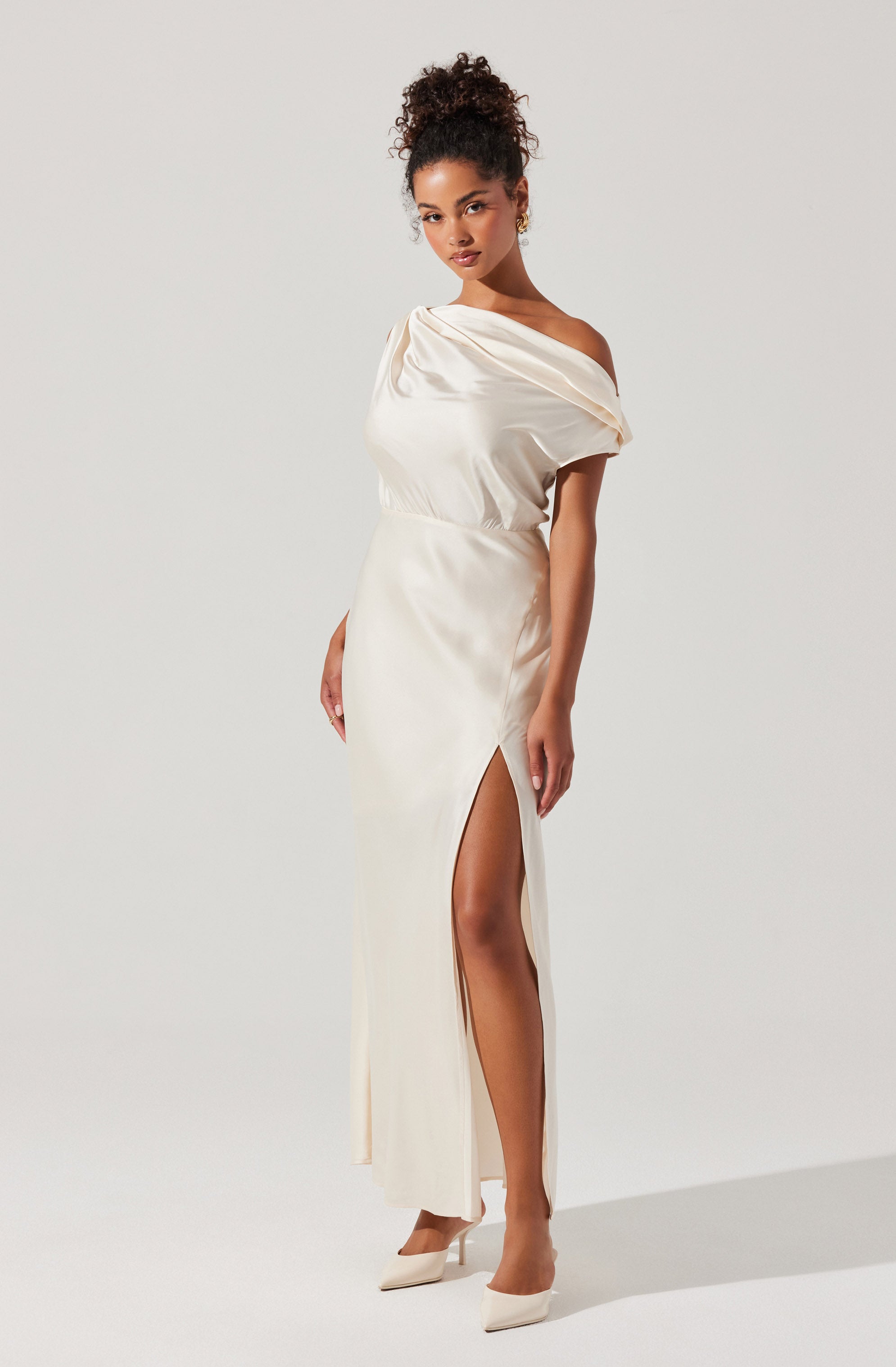 A woman is wearing an elegant off-the-shoulder floor-length gown in a shiny satin fabric. The dress features a draped bodice that falls loosely over her shoulders and a high slit on the side, revealing her leg. She completes the look with a pair of pointed-toe heels in a matching light color.
