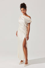 A woman is wearing an elegant off-the-shoulder floor-length gown in a shiny satin fabric. The dress features a draped bodice that falls loosely over her shoulders and a high slit on the side, revealing her leg. She completes the look with a pair of pointed-toe heels in a matching light color.