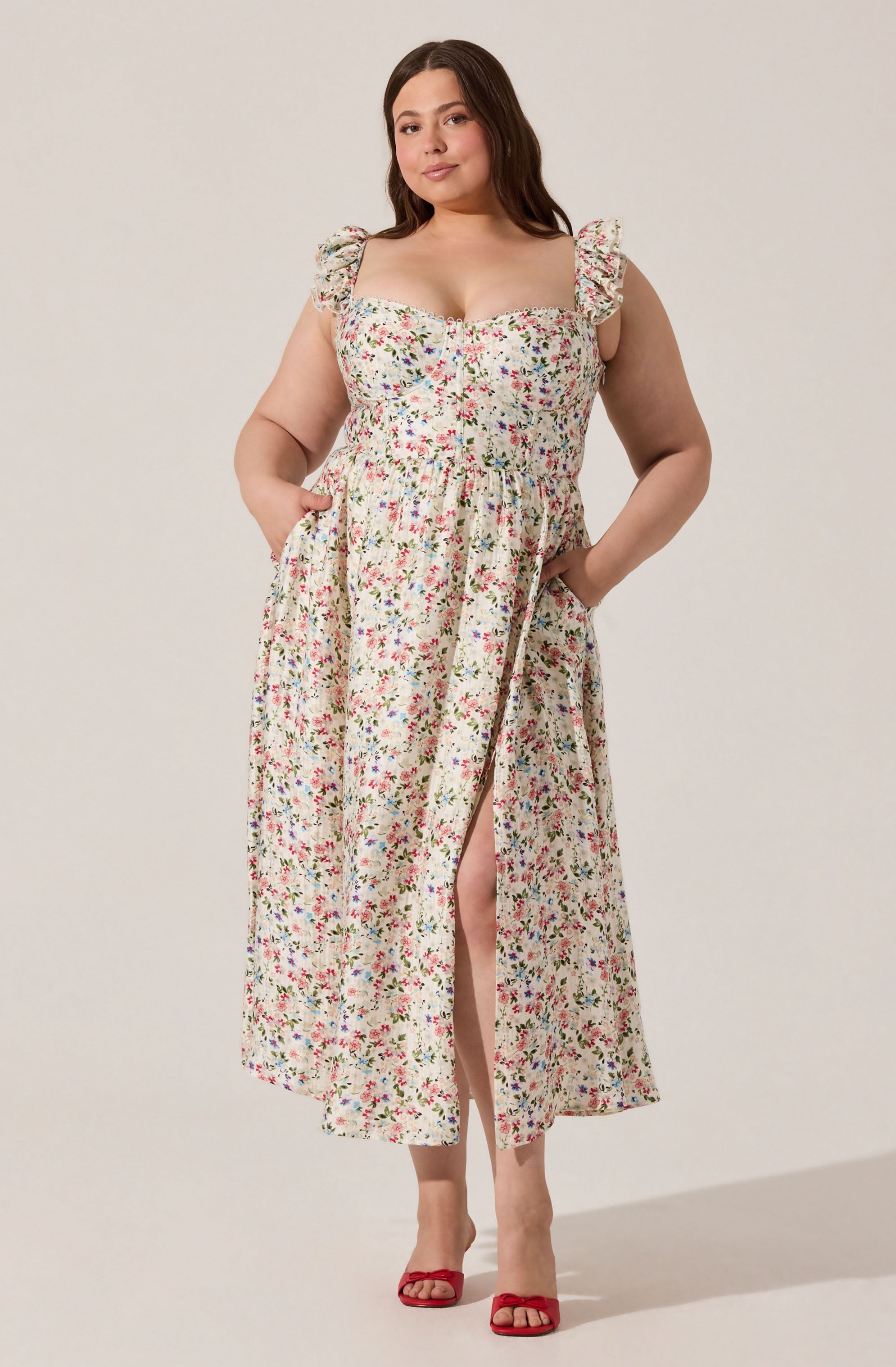 A woman is wearing a sleeveless, ankle-length dress with a floral print pattern. The dress has ruffled shoulder straps, a square neckline, and a gathered waist. She is also wearing red high-heeled sandals.