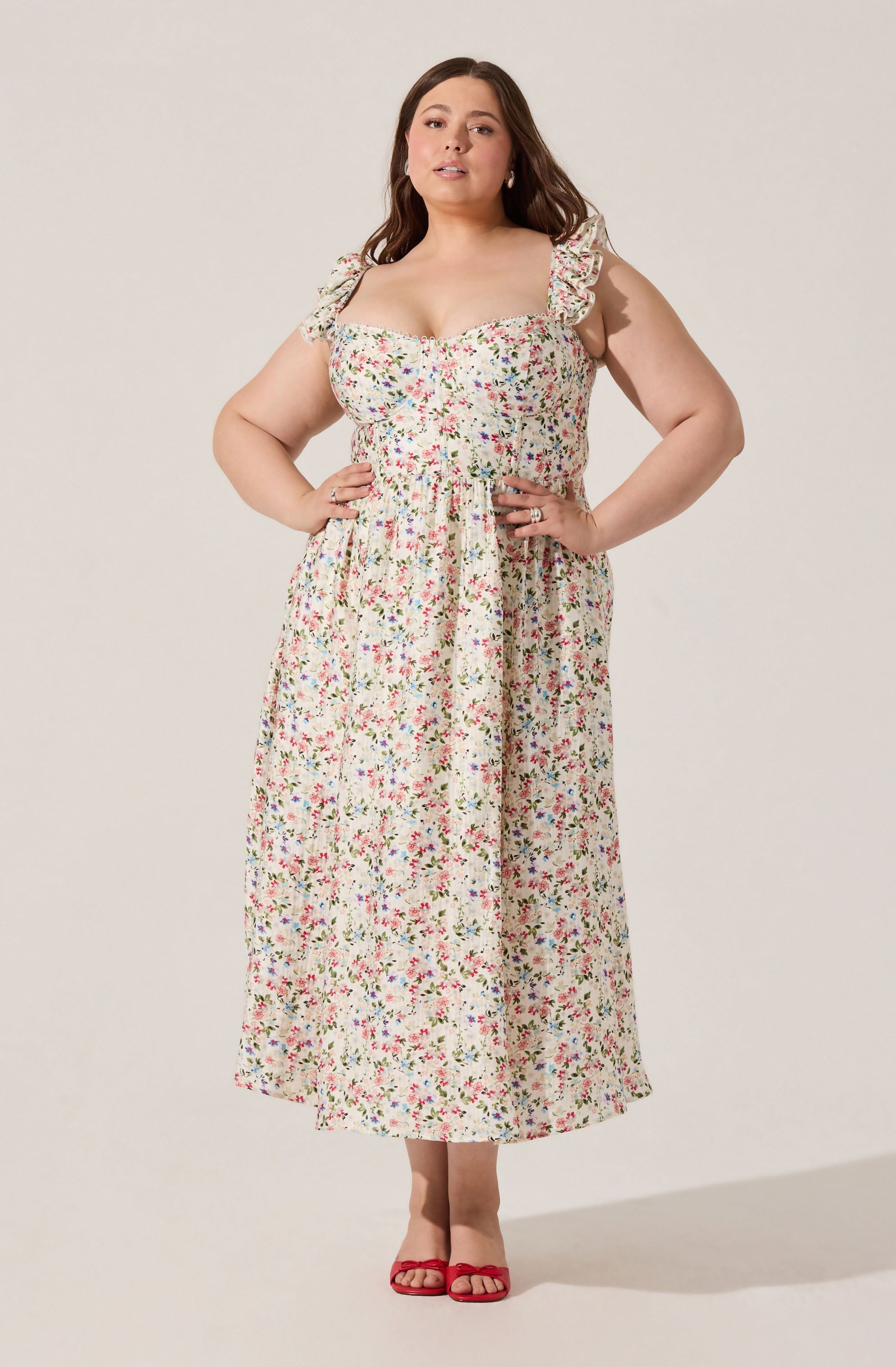 A woman is wearing a sleeveless, long floral dress with ruffled shoulder straps. The dress has a fitted bodice and a slightly gathered waist, flowing down to her ankles. She is also wearing red open-toe sandals.