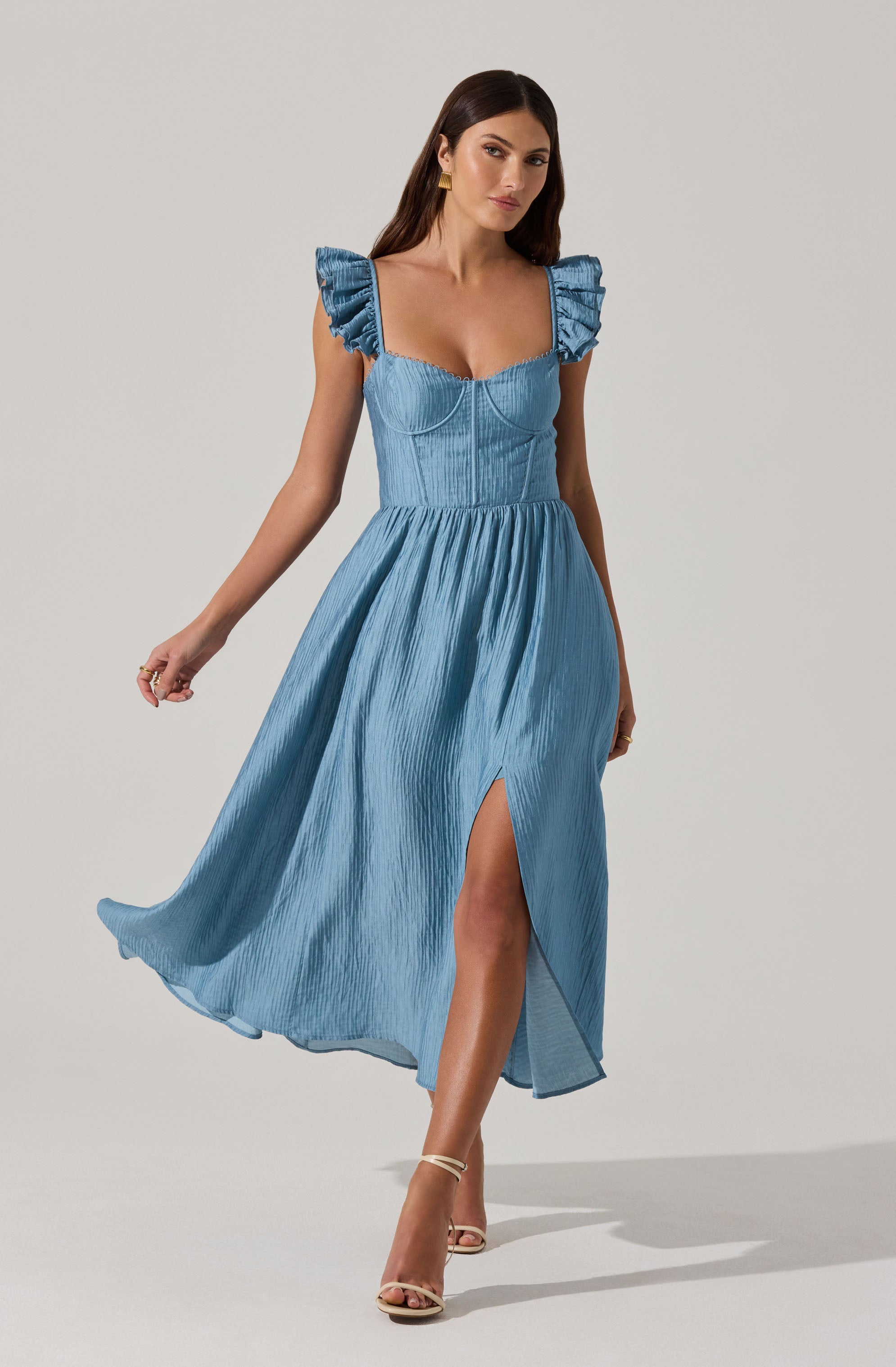 A woman is wearing a light blue, sleeveless dress with ruffled shoulder straps and a fitted bodice. The dress has a high waistline and a flowing, pleated skirt with a thigh-high slit. She is also wearing beige high-heeled sandals.