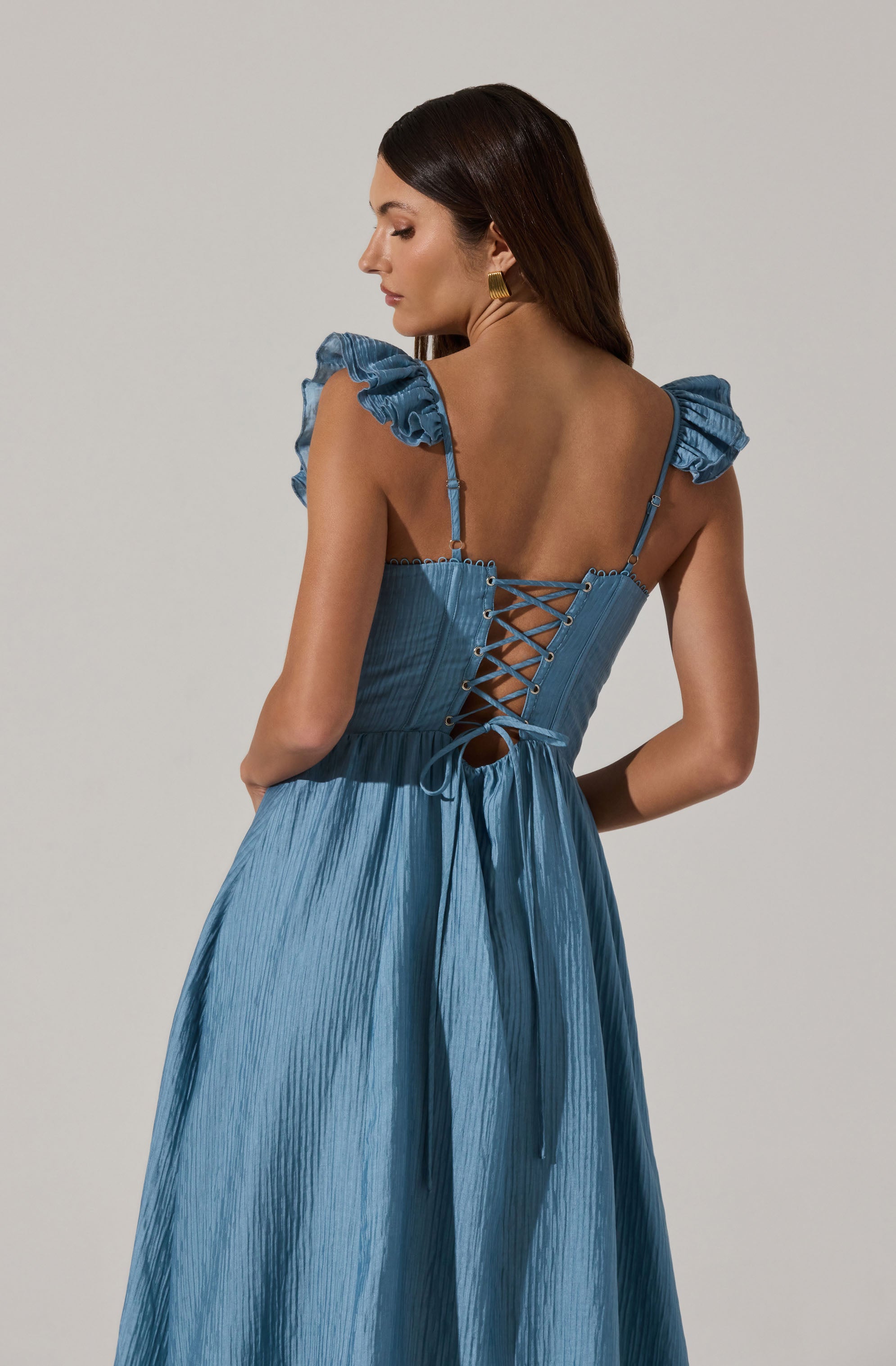 The dress is a light blue, shiny fabric with a pleated texture. It features ruffled shoulder straps and a lace-up back with crisscrossing ties. The waistline is fitted, and the skirt flows down in a wide, gathered style.