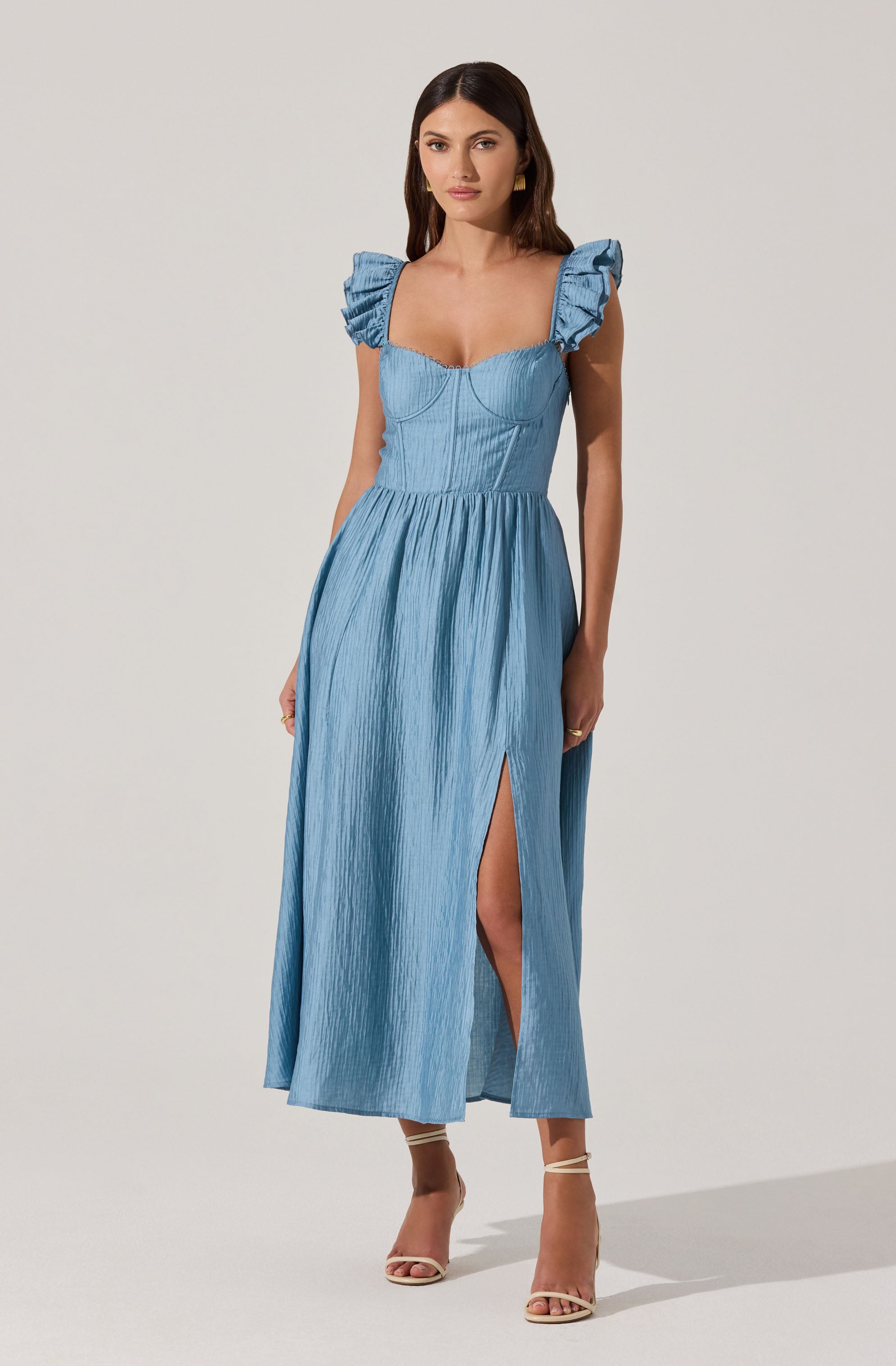 A woman is wearing a light blue maxi dress with textured fabric. The dress features ruffled cap sleeves, a fitted bust with seam details, and a high slit on the left side. She is also wearing nude strappy heels.