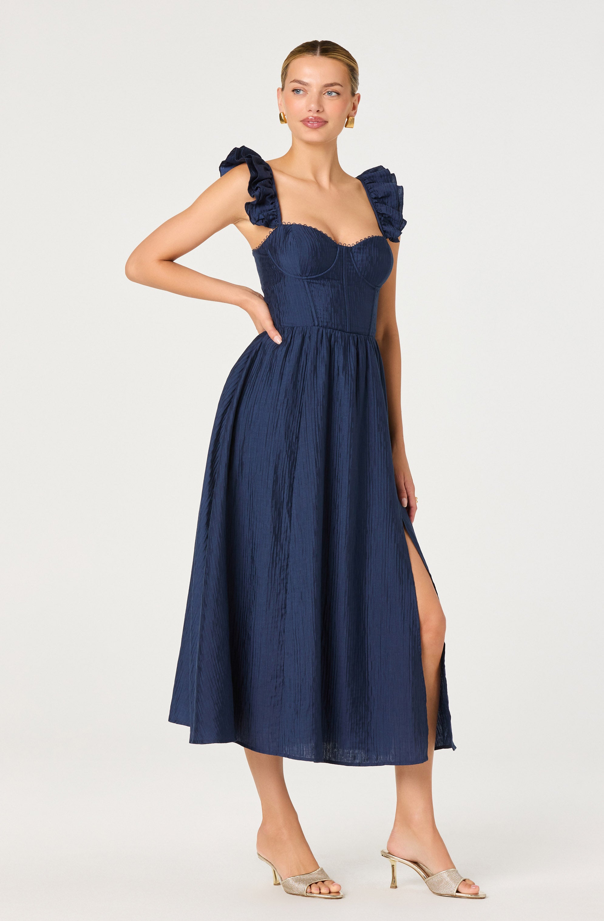 A woman is wearing a dark blue, sleeveless, maxi-length dress with ruffled shoulder straps and a fitted bodice. The dress has a textured, shiny fabric and features a high slit on one side. She is also wearing open-toed heels and gold earrings.