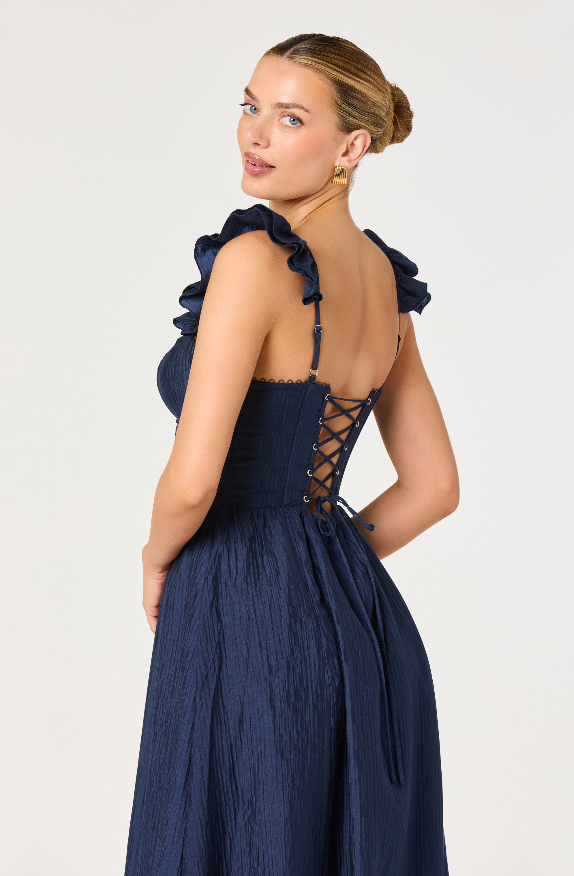 A woman is wearing a navy blue dress with ruffled shoulder straps and a fitted bodice. The back of the dress features a lace-up design with thin straps that tie at the waist. The fabric has a textured, slightly shiny appearance.
