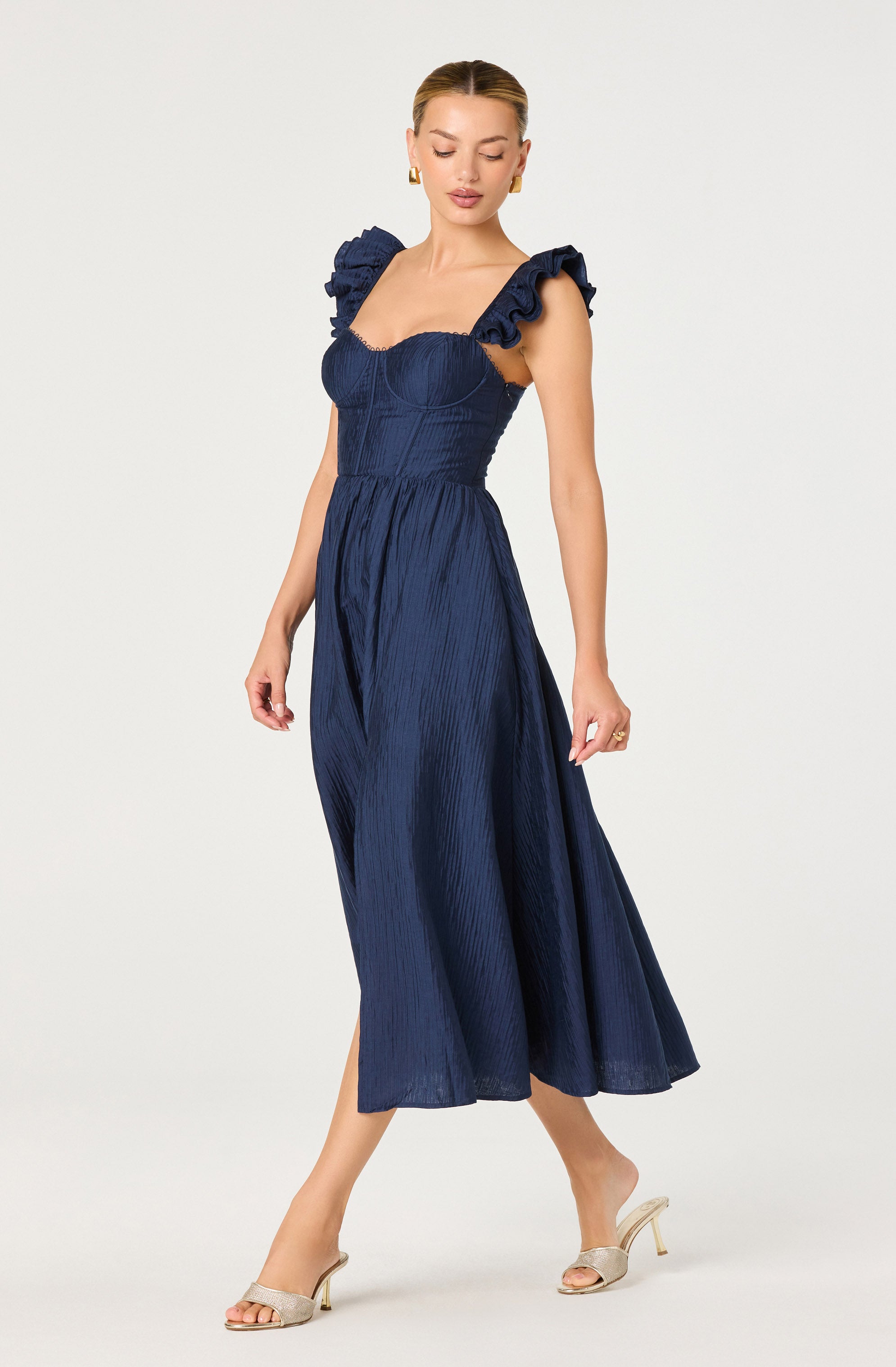 A woman is wearing a navy blue dress with ruffled shoulder straps and a fitted bodice that features vertical textured fabric. The dress has a high waistline and a flowing, ankle-length skirt with a subtle slit. She is also wearing nude-colored open-toe high heels.