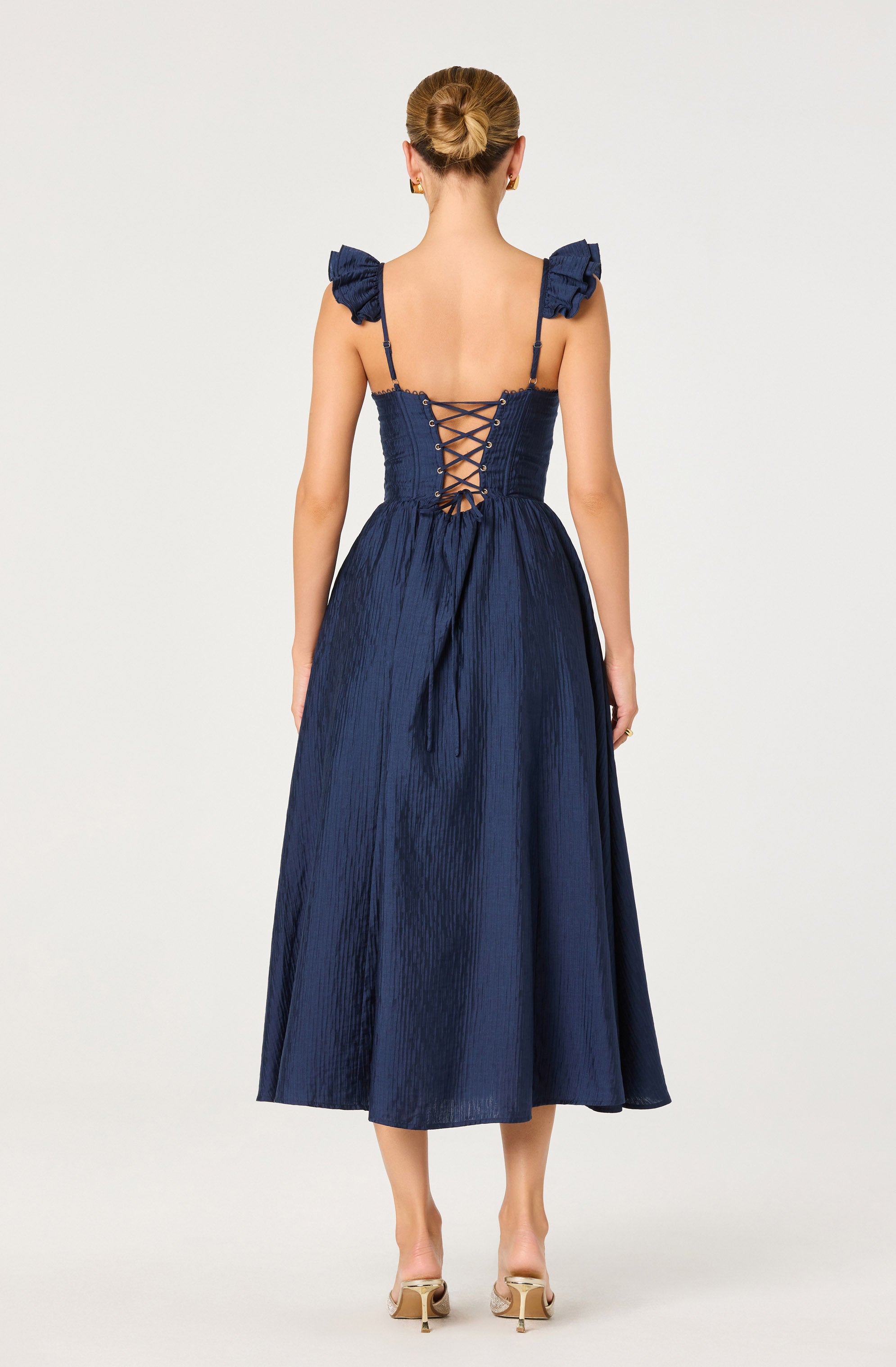 A woman is wearing a navy blue dress featuring a fitted, textured bodice with ruffled shoulder straps. The back of the dress has a corset-style lace-up detail, creating a crisscross pattern. The skirt is long, flowing, and has a shiny, crinkled fabric appearance. She is also wearing nude-colored high heels with a buckle detail.