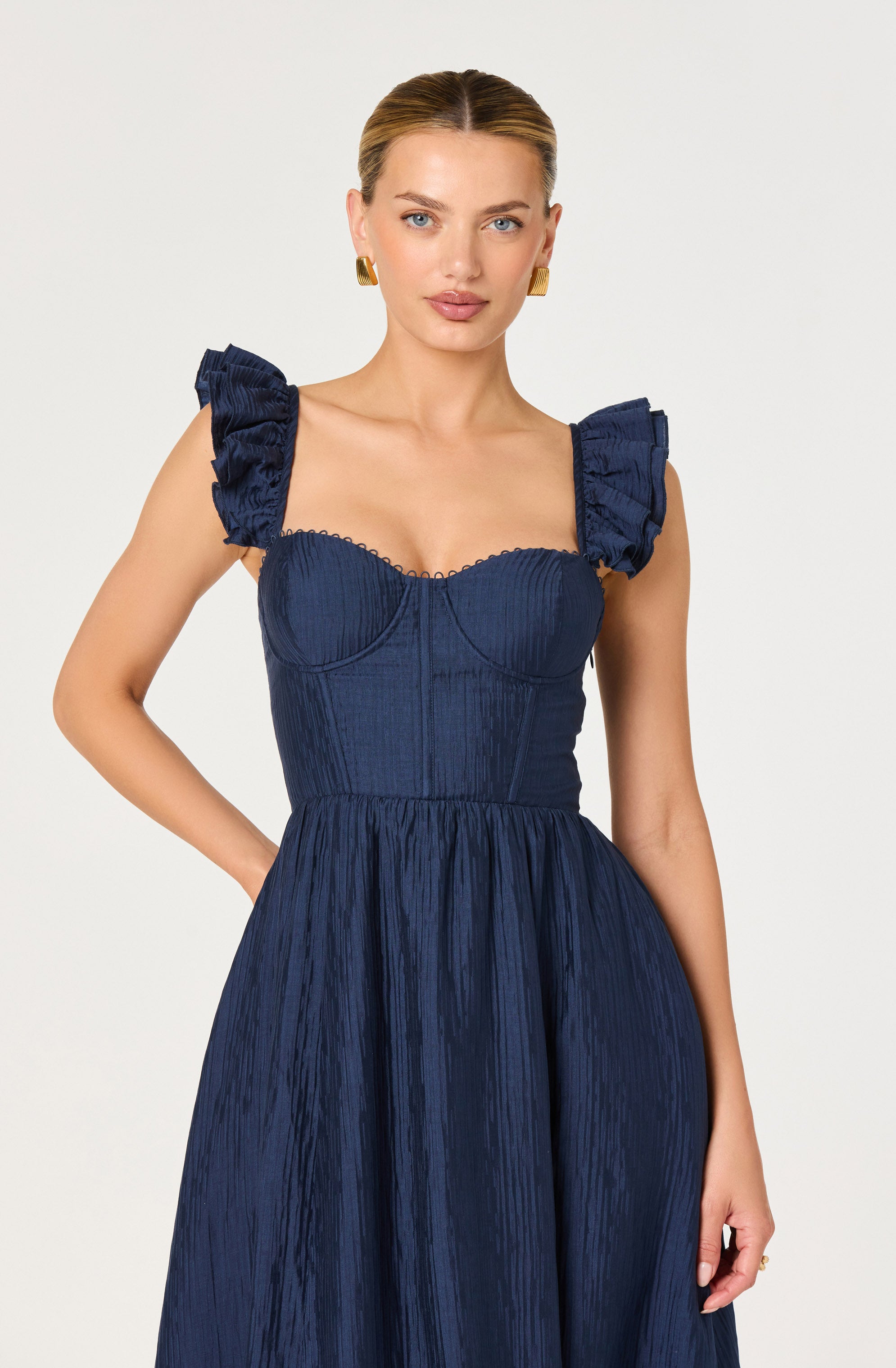 A woman is wearing a dark blue dress with puffed, ruffled shoulder straps. The dress features a fitted, structured bodice with visible vertical seams and a textured, pleated fabric. The skirt is full and gathered, flowing from the waist down in the same textured material. The overall style of the dress is elegant and fashionable.
