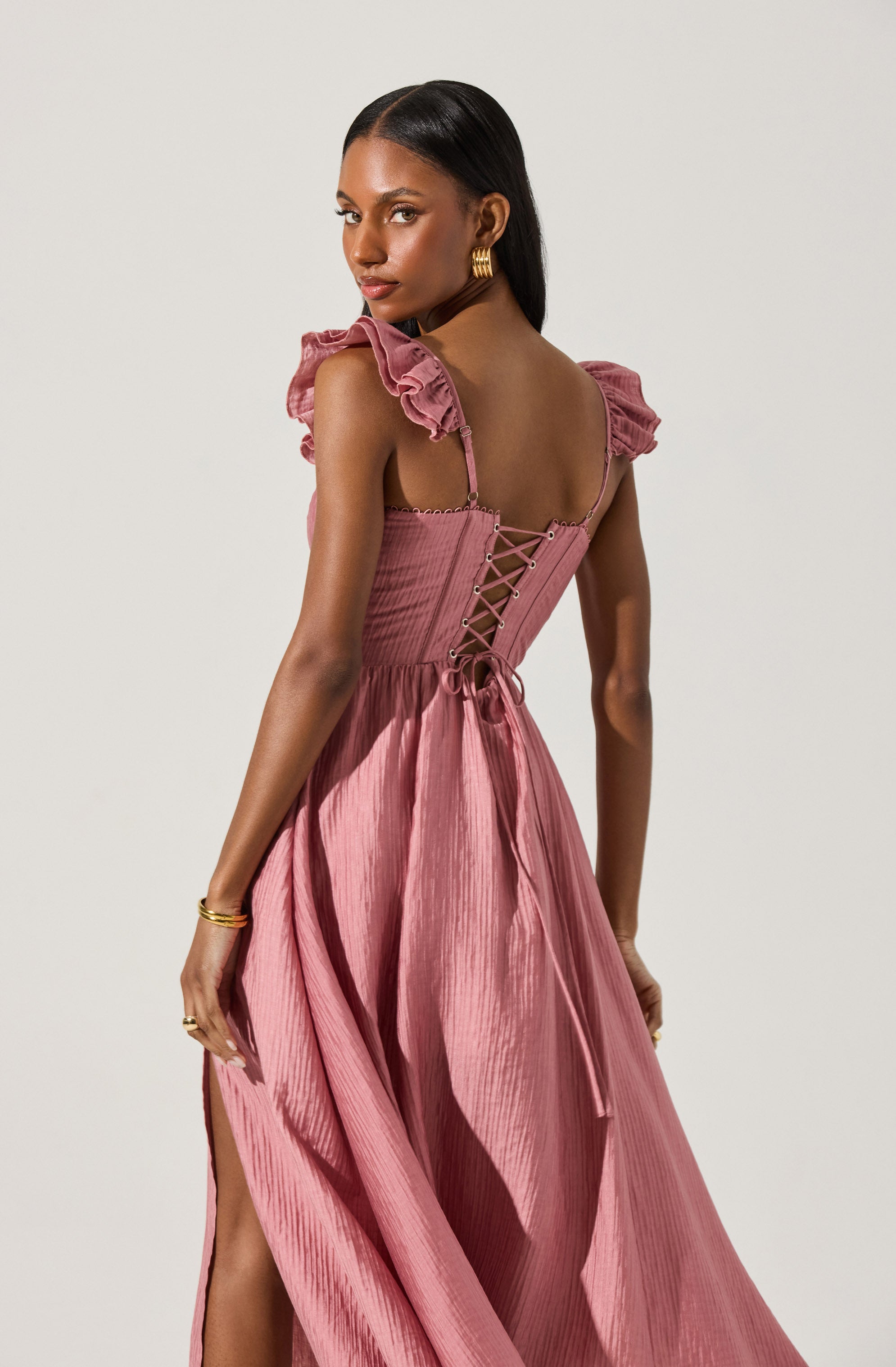 A woman is wearing a pink dress with a fitted bodice that features a lace-up back detail. The dress has ruffled, shoulder straps and a flowy, textured skirt with a slit on one side. She accessorizes with gold hoop earrings and holds her hand gracefully at her side.