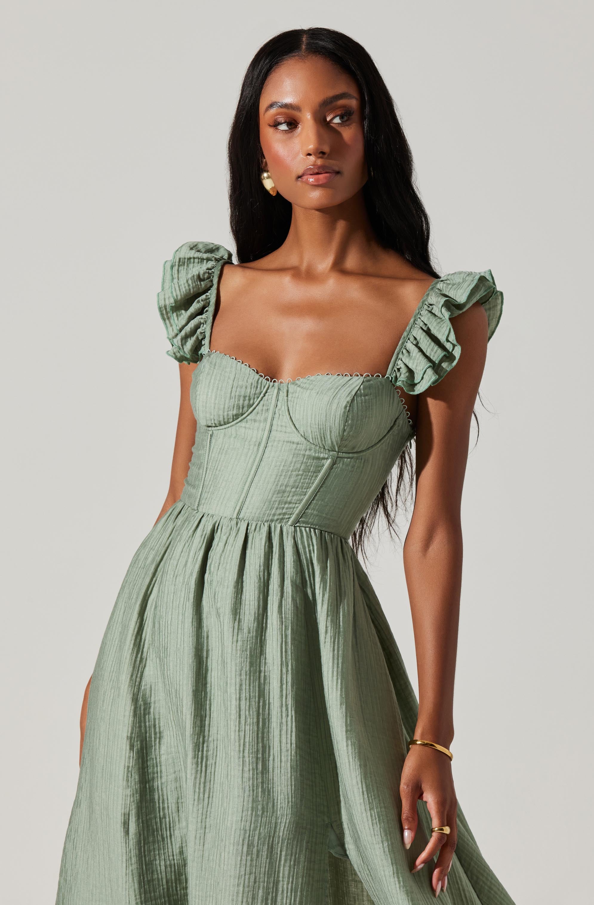 A woman is wearing a light green dress with puffed, ruffled sleeves. The dress has a fitted bodice with visible vertical and horizontal seams, and a full, gathered skirt. The fabric has a slightly textured, shiny appearance. She is accessorized with pearl earrings, a gold bracelet on her right wrist, and gold rings on her fingers.