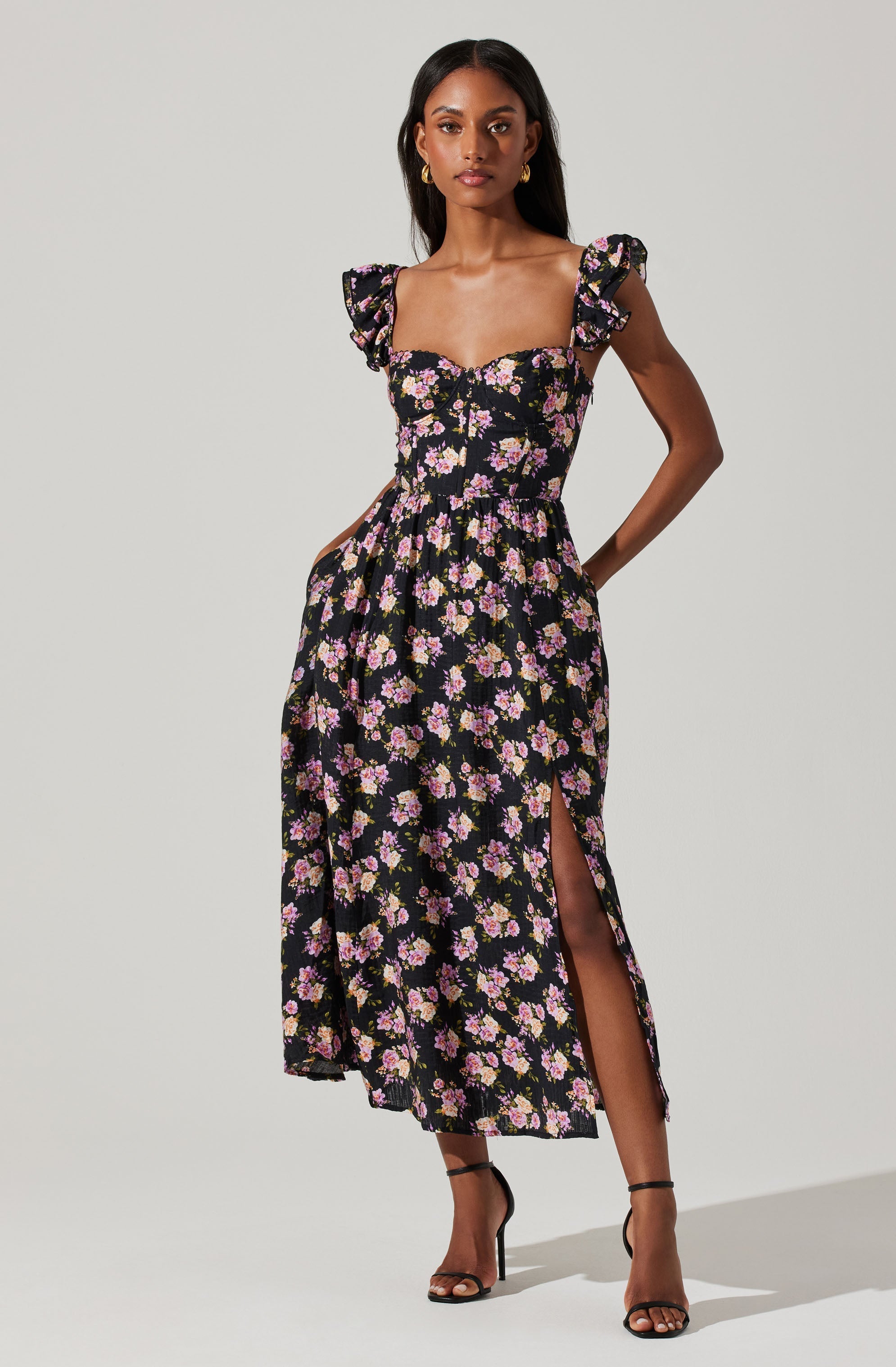 A woman is wearing a black maxi dress with a floral pattern of pink, purple, and white flowers. The dress features ruffled cap sleeves, a fitted bodice with a sweetheart neckline, and a slit on the side. She is also wearing black high-heeled sandals and gold hoop earrings.
