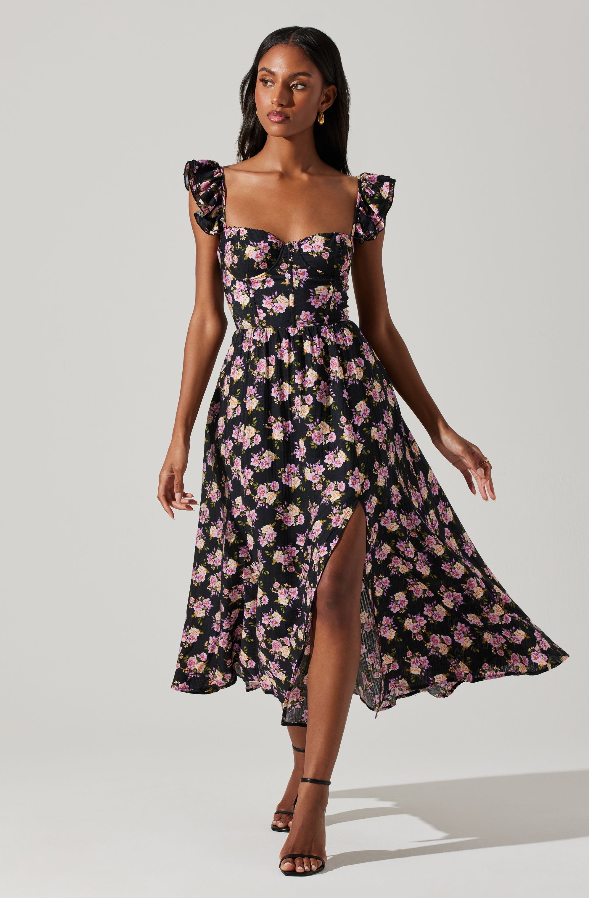 A woman wearing a black dress with a pink, purple, and yellow floral pattern. The dress features a fitted bodice with a sweetheart neckline, ruffled shoulder straps, and a high slit on one side. She is also wearing black high-heeled sandals.