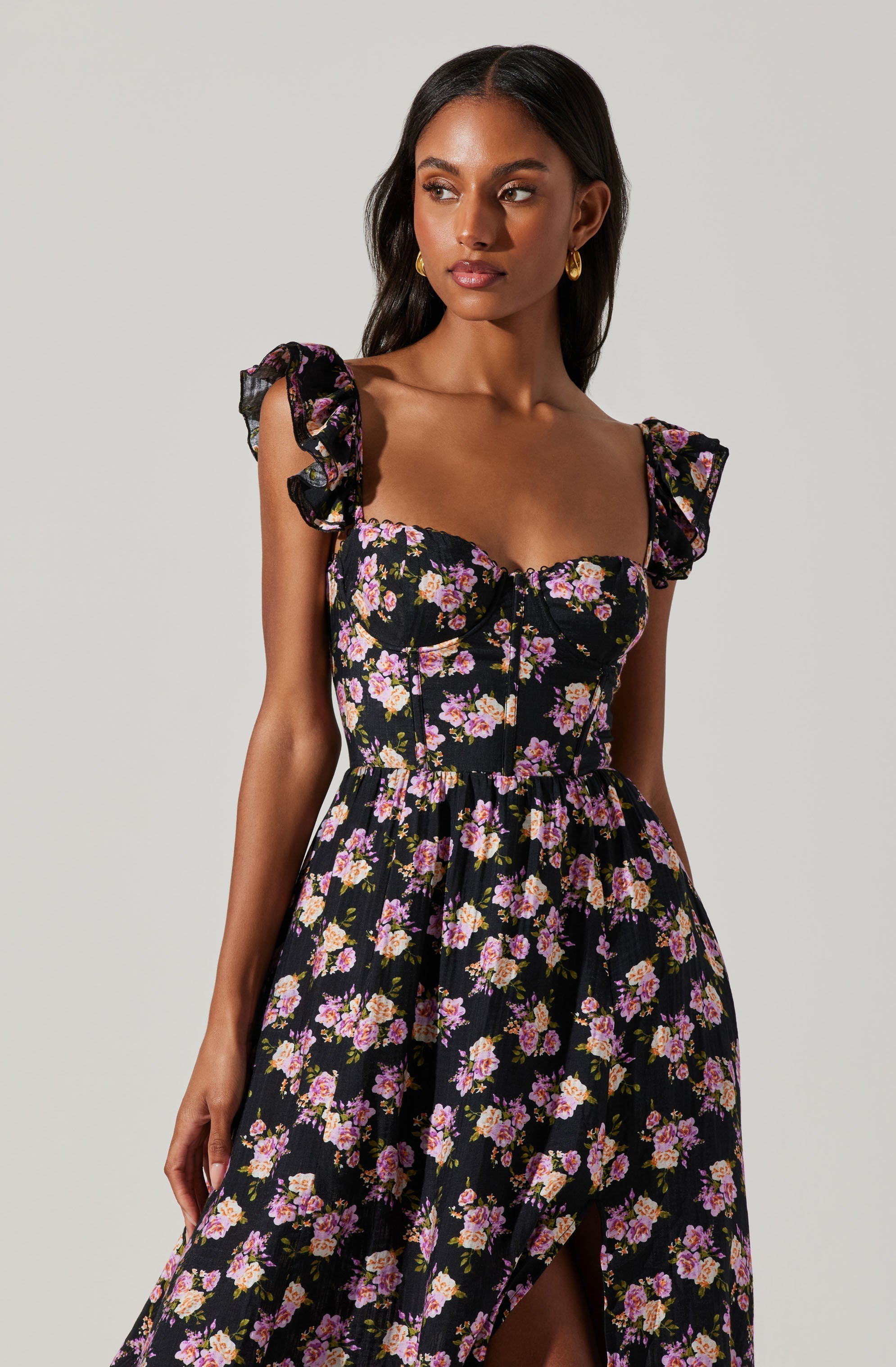 A woman is wearing a black dress with a floral pattern featuring pink, purple, and white flowers. The dress has short, ruffled sleeves and a fitted bodice with a zipper detail in the front. The skirt is flowy with a slit at the front.