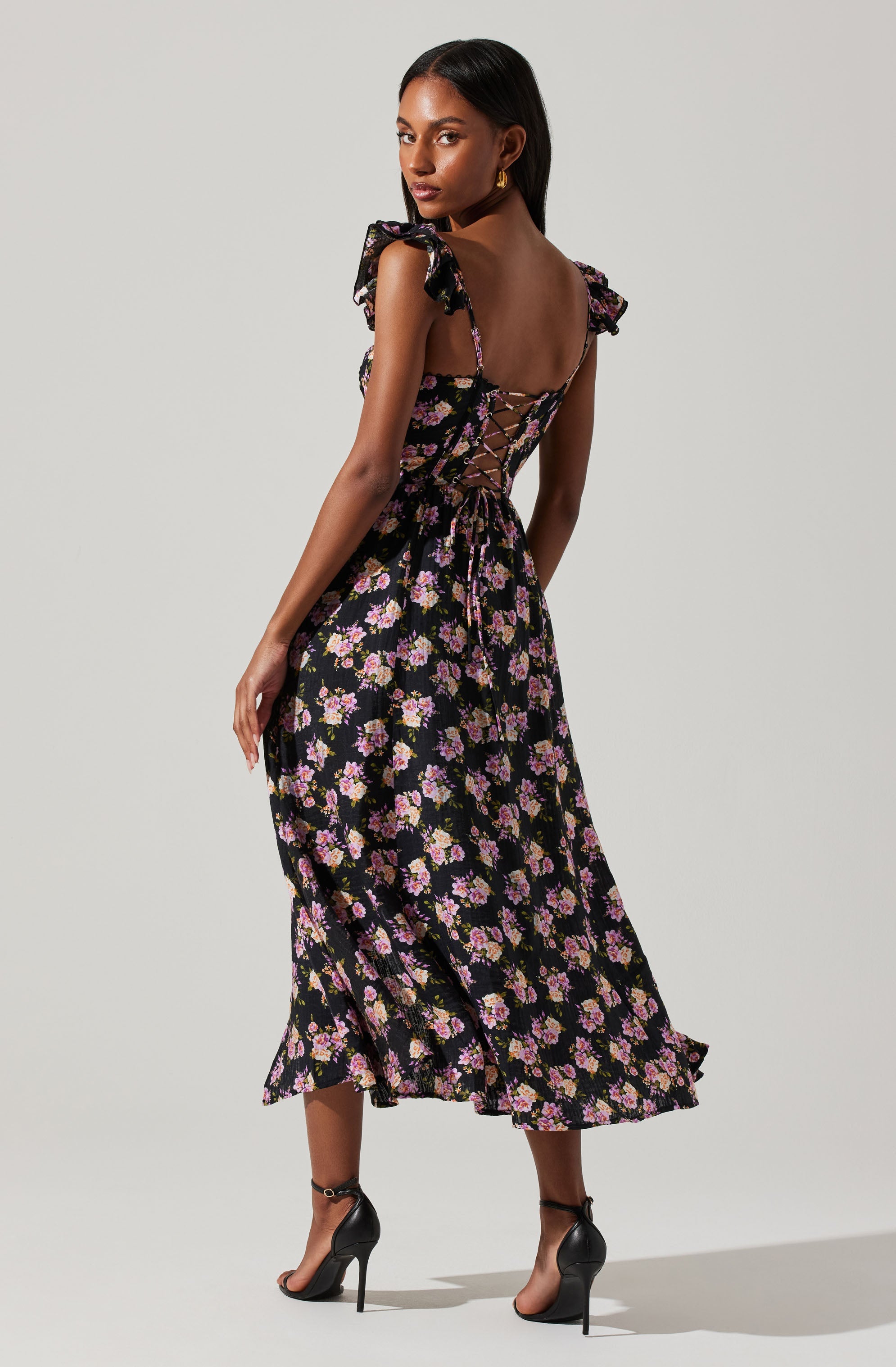 A woman is wearing a long black dress with a floral pattern of pink, purple, and white flowers. The dress features ruffled shoulder straps, a fitted bodice with a lace-up back, and a flowing skirt that reaches mid-calf. She has on black high-heeled sandals with ankle straps and is accessorized with gold earrings.