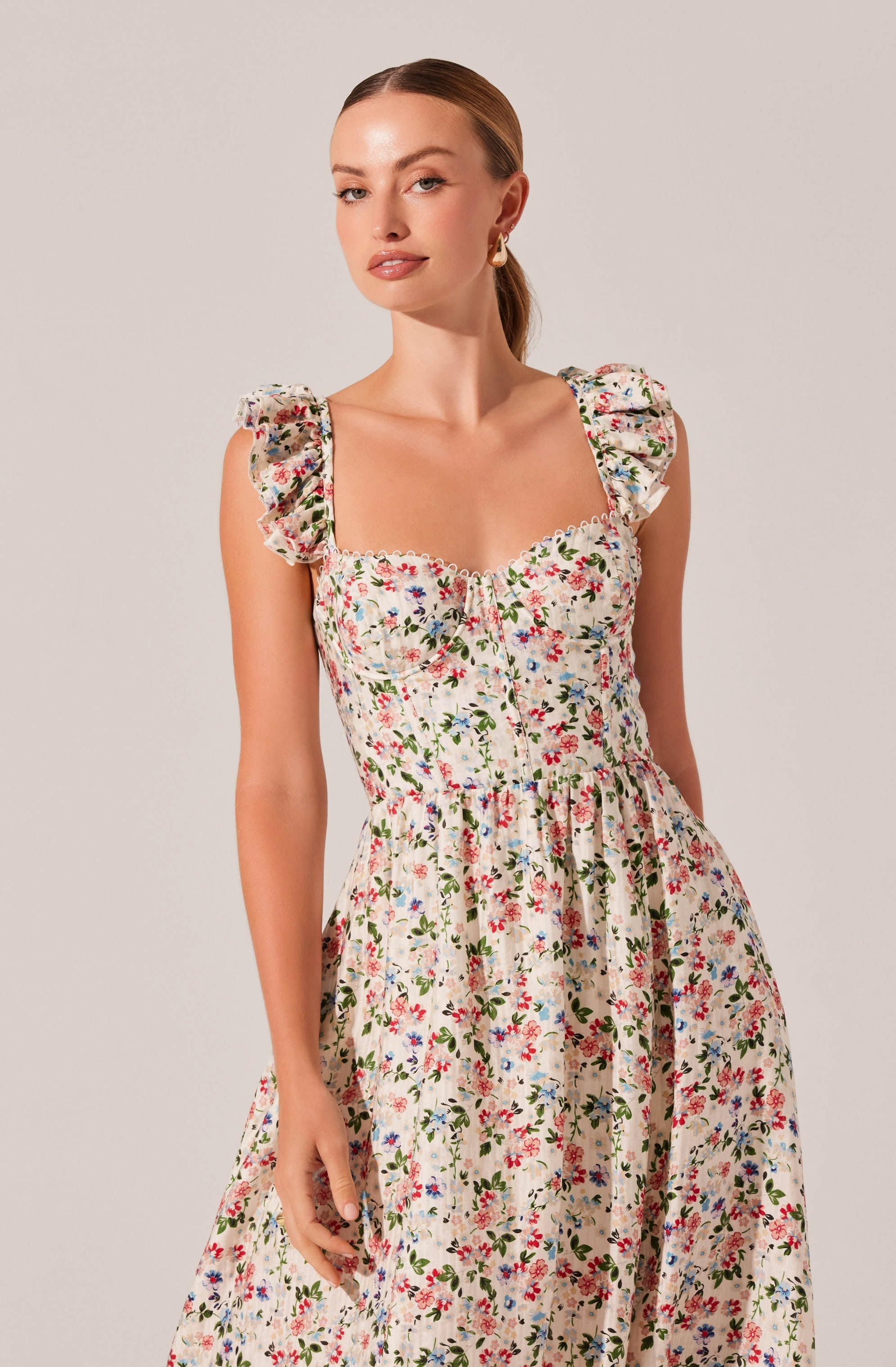 A woman is wearing a sleeveless, floral dress with ruffled shoulder straps, featuring a pattern of small, colorful flowers on a light background. The dress has a fitted bodice with a square neckline and a gathered waist that flows into a full skirt.