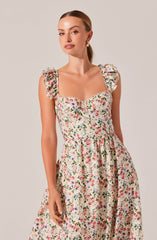 A woman is wearing a sleeveless, floral dress with ruffled shoulder straps, featuring a pattern of small, colorful flowers on a light background. The dress has a fitted bodice with a square neckline and a gathered waist that flows into a full skirt.