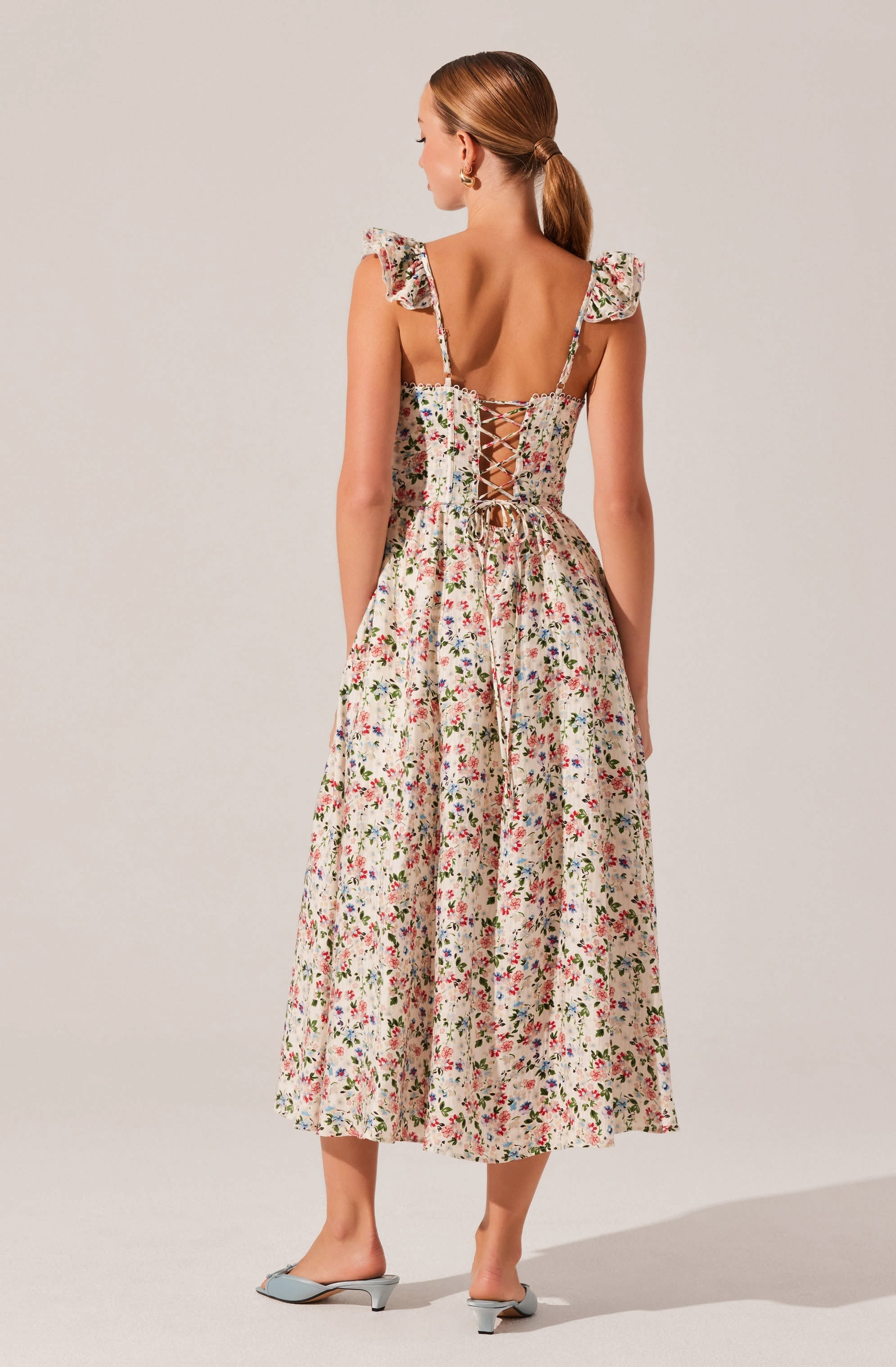 A woman is wearing a sleeveless, floral print dress with ruffled shoulder straps. The back of the dress features a lace-up detail with a crisscross pattern. She is also wearing light-colored, open-toed heels with a small heel.