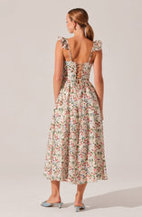 A woman is wearing a sleeveless, floral print dress with ruffled shoulder straps. The back of the dress features a lace-up detail with a crisscross pattern. She is also wearing light-colored, open-toed heels with a small heel.