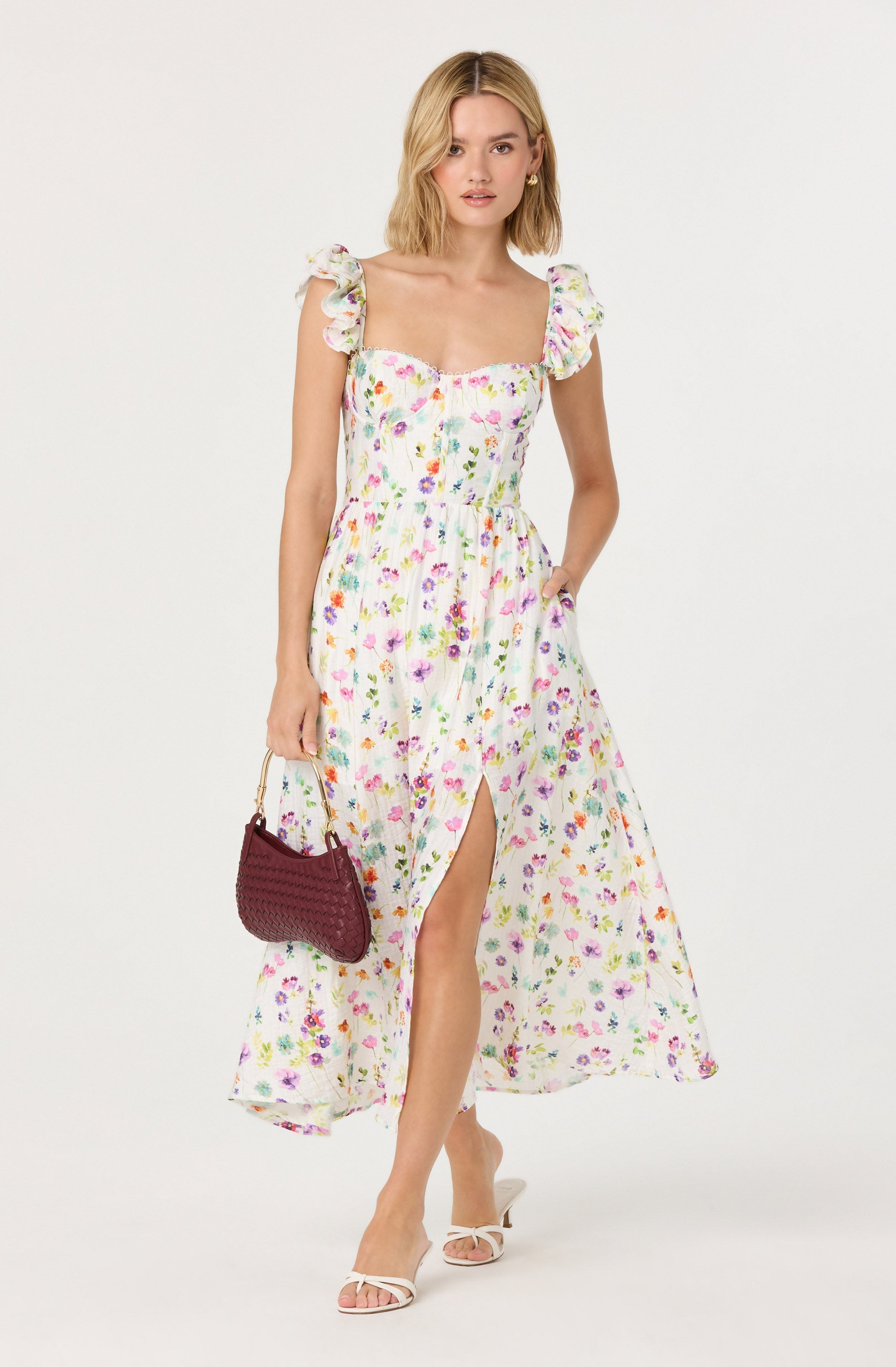 A woman is wearing a long, sleeveless, floral dress with ruffled shoulder straps. The dress has a sweetheart neckline, a fitted waist, and a front slit. She is holding a dark red woven handbag with a gold handle and is wearing white, open-toe high-heeled shoes.